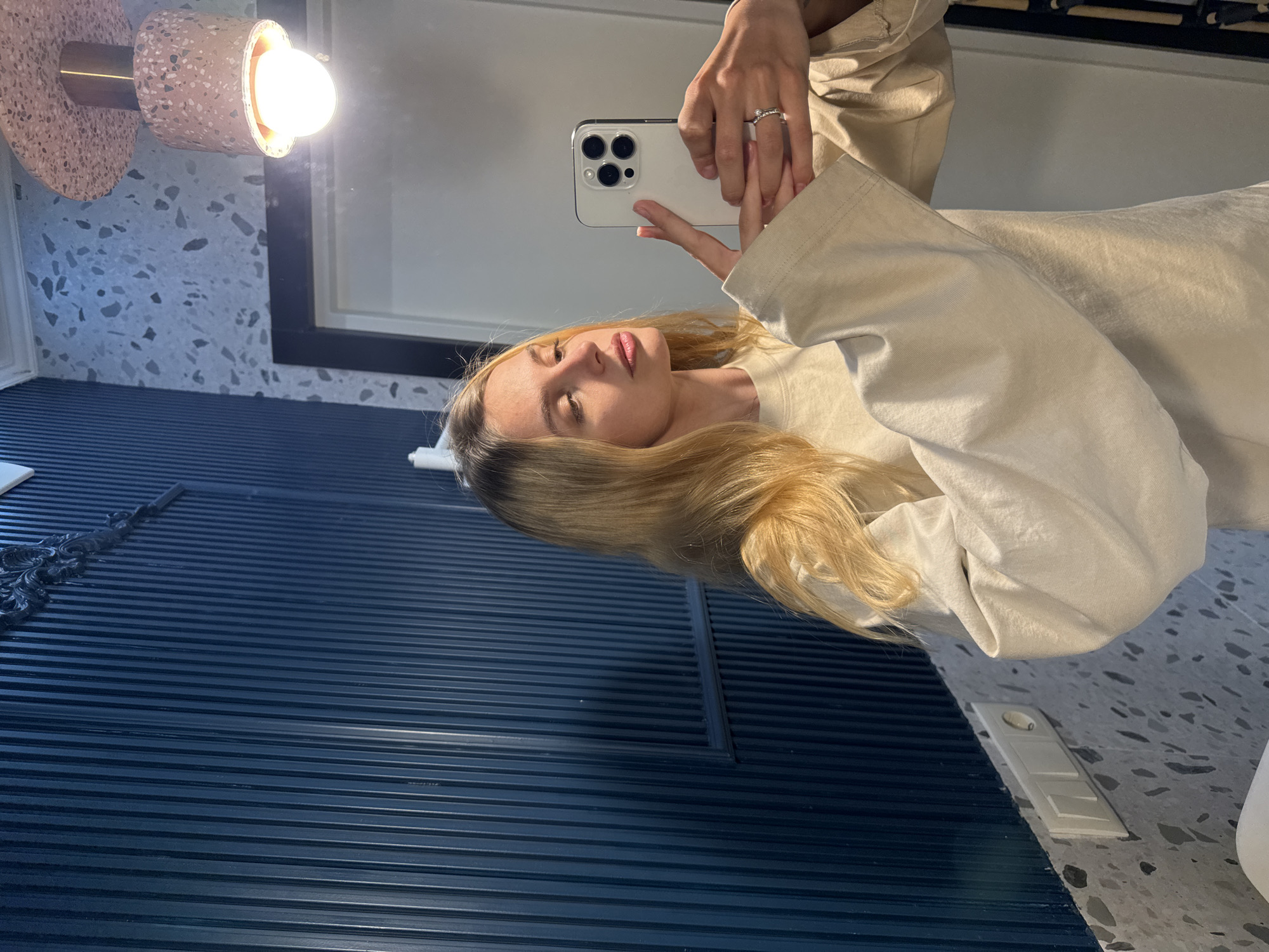 A woman with long blonde hair is taking a selfie in a bathroom mirror. She is wearing a light beige, long-sleeved top and holding a white smartphone with multiple camera lenses. A ring is visible on her finger. In the background, there is a blue slatted wall, a light fixture with a speckled pink and gold design, and a white speckled countertop. The bathroom appears to be in an unknown city.