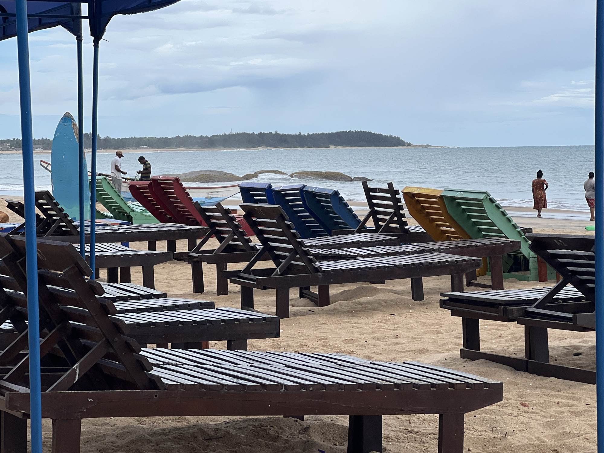 The image captures a beach scene in Arugam Bay, Sri Lanka, under an overcast sky. The foreground is dominated by multiple rows of unoccupied wooden sun loungers, oriented towards the ocean. These loungers are painted in various colors including green, red, blue, and yellow. In the background, two people stand near a blue surfboard and what looks like small boats. Further along the beach, two more people are walking near the water's edge. The sea is choppy, and a strip of land is visible in the distance.