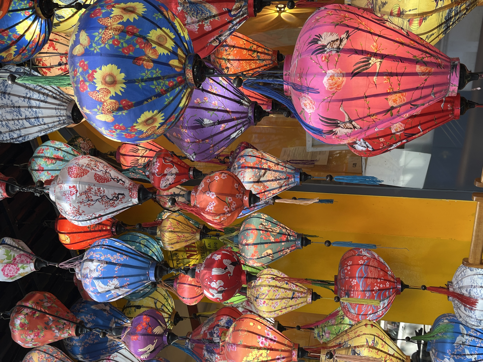 The image depicts a vibrant collection of colorful lanterns hanging from a ceiling in what appears to be a shop or market in Đà Nẵng, Vietnam. The lanterns are made of silk or fabric, with various floral and bird patterns. Dominant colors include blue, pink, purple, red, orange, and yellow. The lanterns are suspended in rows and are in close proximity to each other, creating a dense and visually stimulating display. The background is a flat yellow surface with other decorative elements.