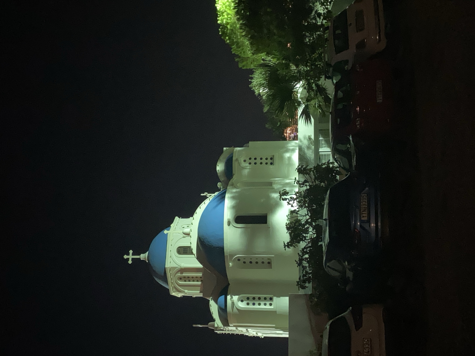 In the city of Thira Municipal Unit, Greece, a white building with blue domes and a cross on top, appears to be a church, is lit up at night. The building is ornate, with arched windows and multiple levels. In front of the building, several cars are parked. The scene is dark, with the exception of the illuminated building and streetlights, casting shadows on the ground. There are trees in the background, some of which are illuminated by the building's lights. The overall impression is one of a quiet, serene night in a picturesque location.