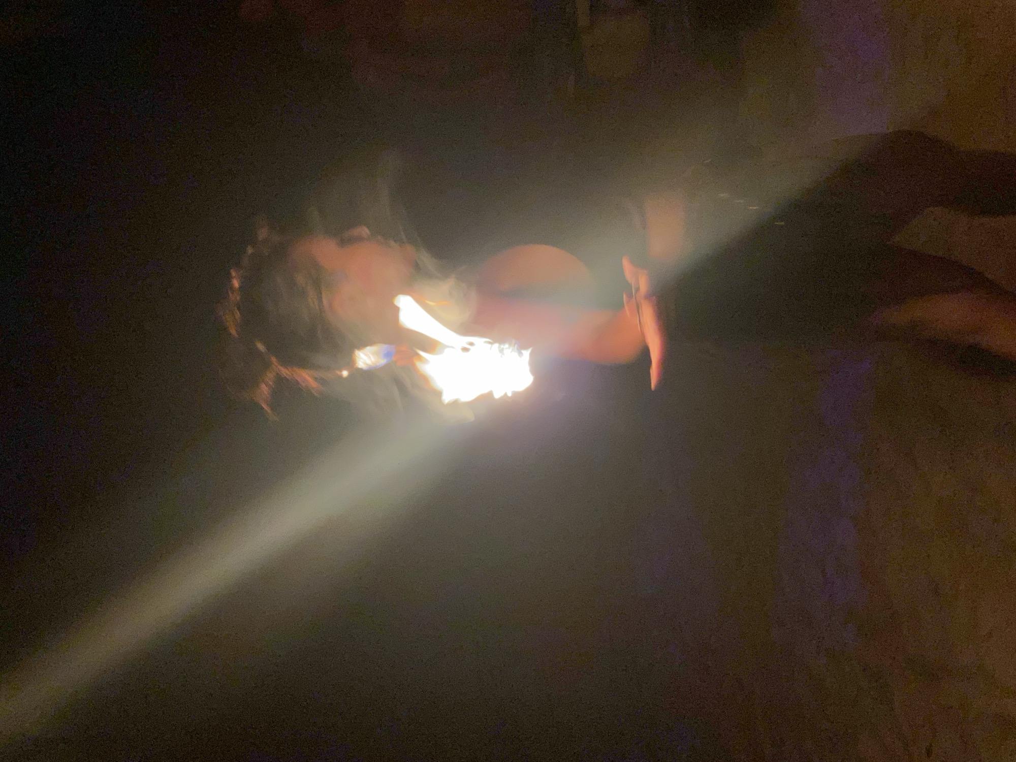 A blurry, low-light photograph appears to depict a person in Baan Sri Thanu, Thailand, likely performing a fire-breathing act. The person, facing left, has their head tilted back as a burst of flames erupts from their mouth. The light from the fire illuminates their face and surrounding hair, which appears somewhat unkempt. Their left arm is visible, held up in the air with a hand near the flames, possibly holding an instrument or fuel source. The background is dark and indistinct, with some shadows and a few blurry shapes. A light beam is coming from the bottom left.