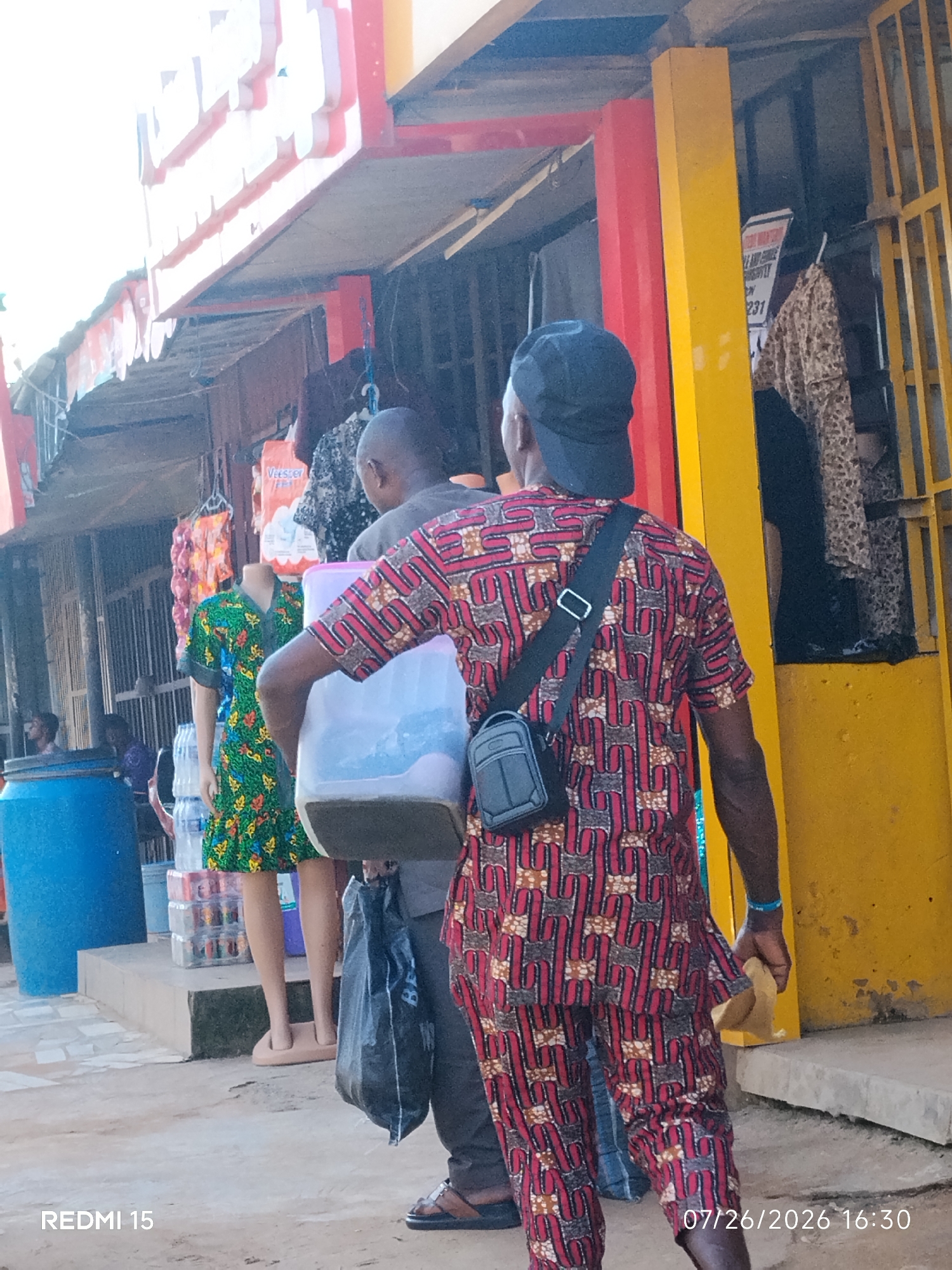 This photograph captures a street scene in Umuode, Nigeria, likely depicting a marketplace or a commercial area. In the foreground, a man wearing a patterned red and black dashiki shirt and pants, a black baseball cap, and a crossbody bag walks away from the camera. He is carrying a large, clear plastic container with a blue lid in his right hand and a black plastic bag in his left. To his left, a blue cylindrical barrel is partially visible. Behind the man, a mannequin dressed in a green and floral patterned short-sleeved dress stands on display. Several stacks of bottled drinks are arranged behind the mannequin, with a white plastic bottle on top of one stack. Further in the background, more clothing items hang on display, and a yellow metal gate is visible, partially obscured by a red and yellow structure. The environment suggests an outdoor commercial setting with stalls or shops lining the street. The lighting indicates it is daytime, with strong sunlight casting shadows. The overall atmosphere appears to be that of a busy commercial area.