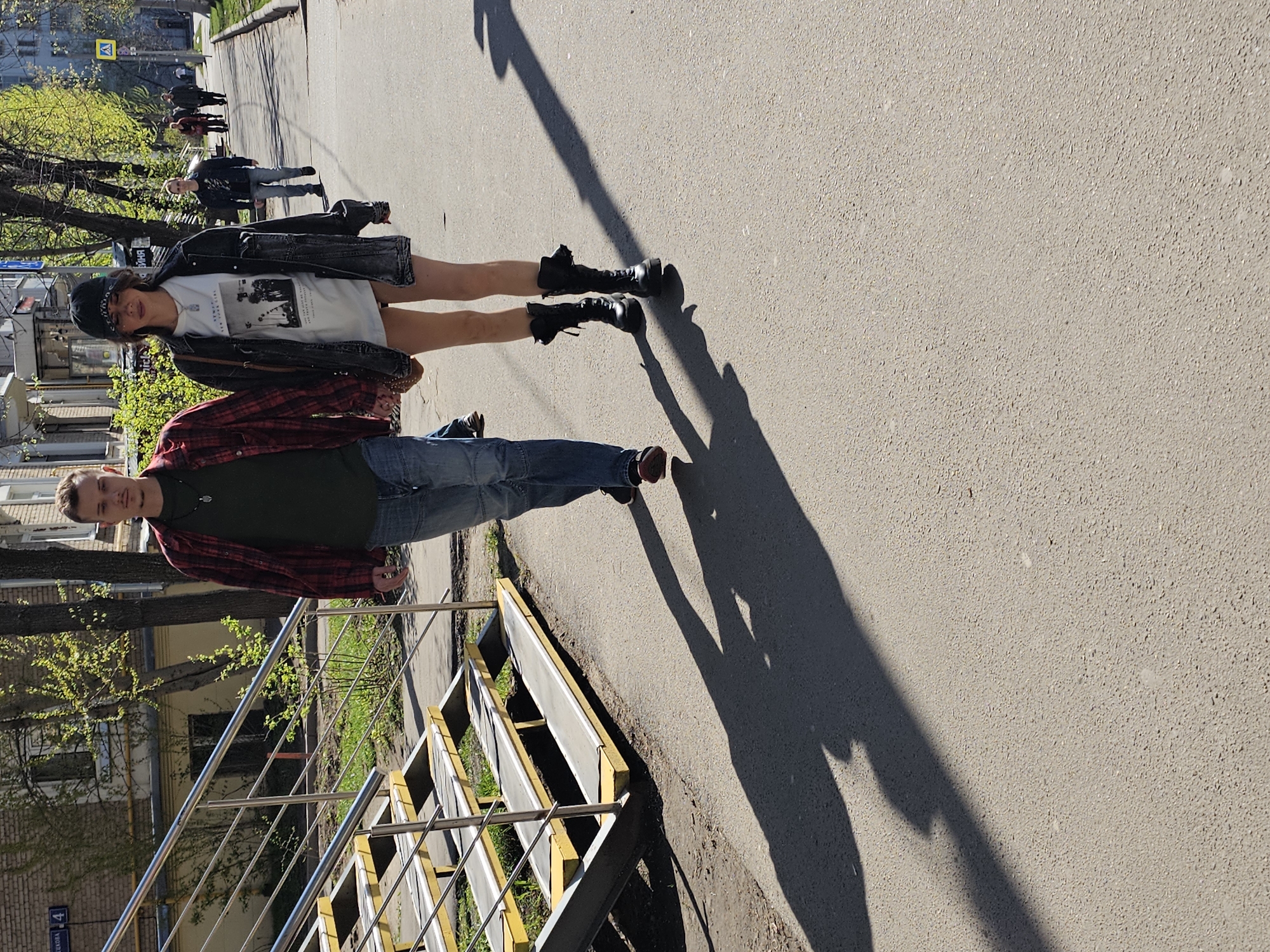 The image shows two individuals on a city street. One person, a young man, is standing near a set of stairs with metal railings and yellow supports. He is wearing jeans, a dark green shirt, and a red plaid overshirt. The other person, a young woman, is standing behind the man. She is wearing black boots, a white T-shirt with a graphic print, and a black jacket. She also has a black cap on her head. The sun is casting a shadow of the pair onto the street. In the background, more people are walking down the sidewalk and buildings and trees can be seen.
The city is identified by the street sign in the image as Khimki, Russia.