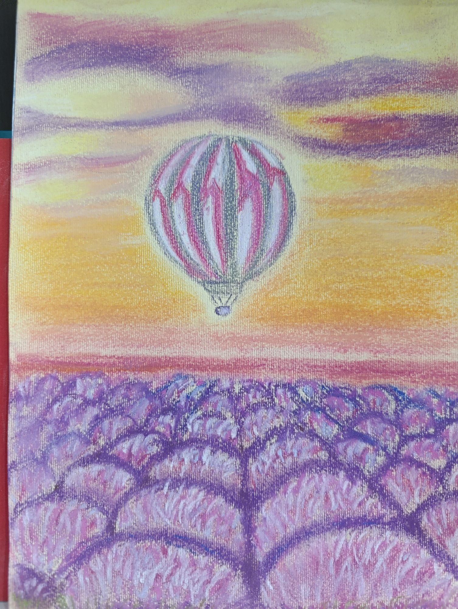 A vibrant artwork depicts a hot air balloon soaring above a field under a multi-hued sky. The upper portion of the image features a sky gradient, transitioning from soft yellow and orange hues at the horizon to patches of purple and pink further up, suggesting either dawn or dusk. A large hot air balloon, centrally positioned, displays vertical stripes of white/light grey and deep pink/magenta, with darker outlines giving it form. A small, dark purple gondola is visible beneath the balloon. Below the horizon line, the lower half of the painting portrays an expansive field. This field is characterized by rich purple and lilac tones, with individual strokes of lighter pink and white suggesting flowering plants, possibly lavender, arranged in distinct rows or clumps. Darker purple lines within the field contribute to a sense of depth and pattern. The entire artwork exhibits a visible canvas texture, indicative of a painting, possibly rendered with pastels or chalks due to the soft, blended appearance of colors and distinct strokes. There are no individuals or specific geographical landmarks identifiable in the scene.