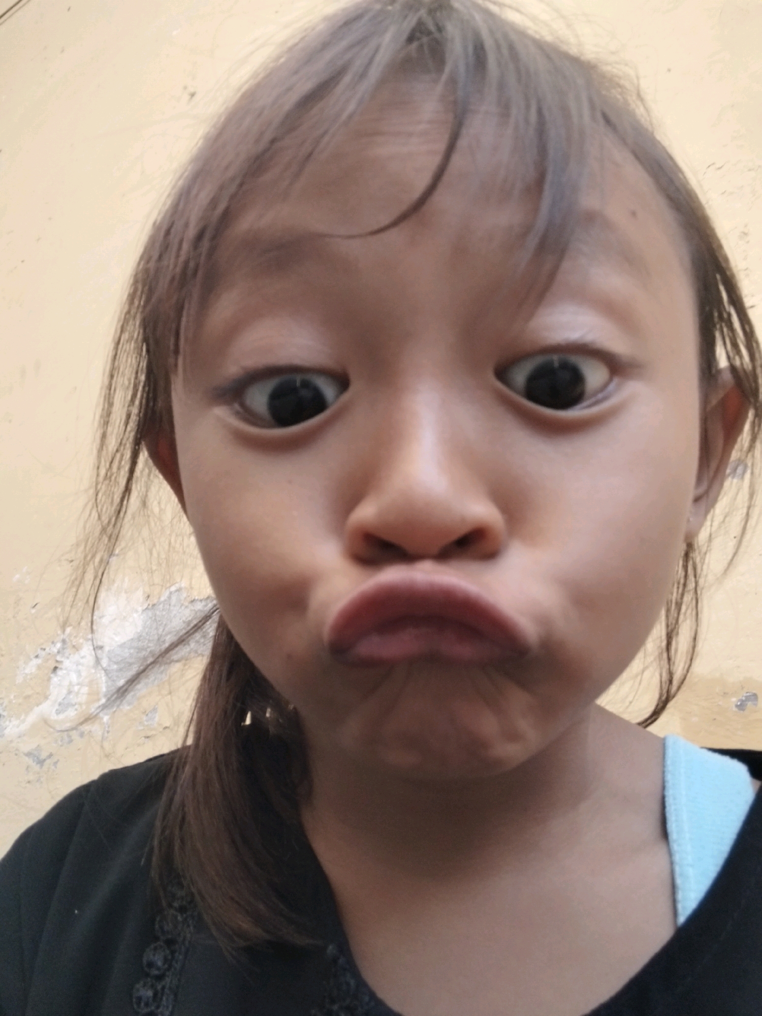 The image is a close-up, high-angle, selfie-style shot of a young girl with her eyes wide and her lips pursed. She has dark hair that is partially obscuring her face. She is wearing a black shirt with a light blue strap visible at the neckline. The background appears to be a textured, light-colored wall. The image was taken in Kepuh Kiriman, Indonesia.