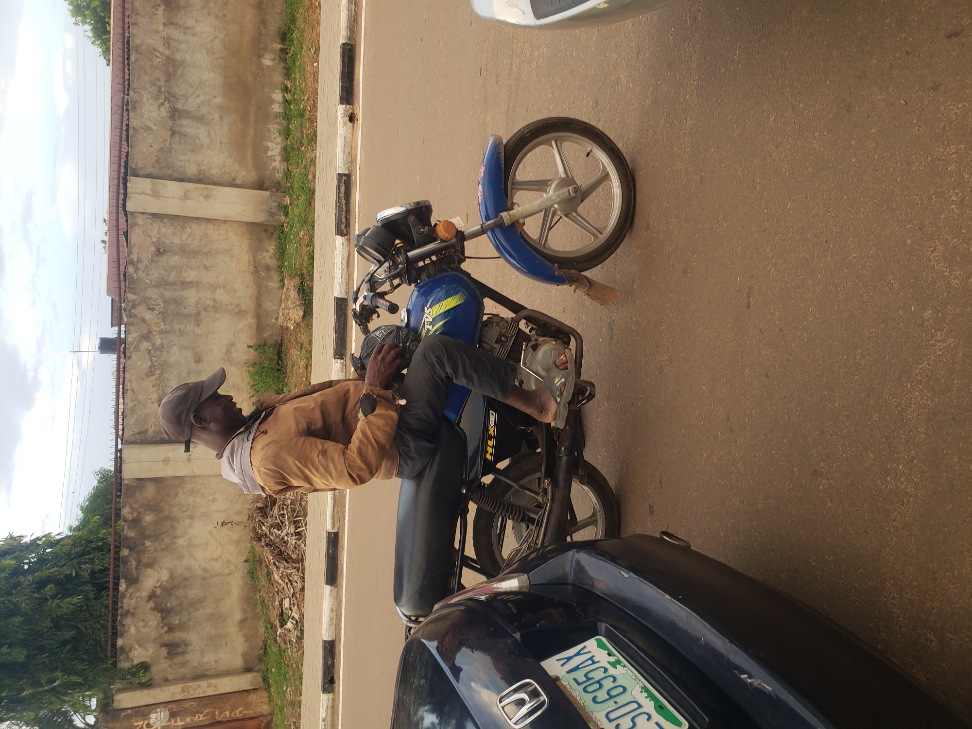 A man wearing a brown jacket and a baseball cap is sitting on a blue motorcycle on a paved road in Narayi, Nigeria. The motorcycle has a black seat and a silver wheel. A dark blue car with a white and green license plate is parked to the left of the motorcycle. In the background, there is a concrete wall and some trees. The sky is cloudy. The man appears to be looking down at his hands, which are resting on the handlebars of the motorcycle.