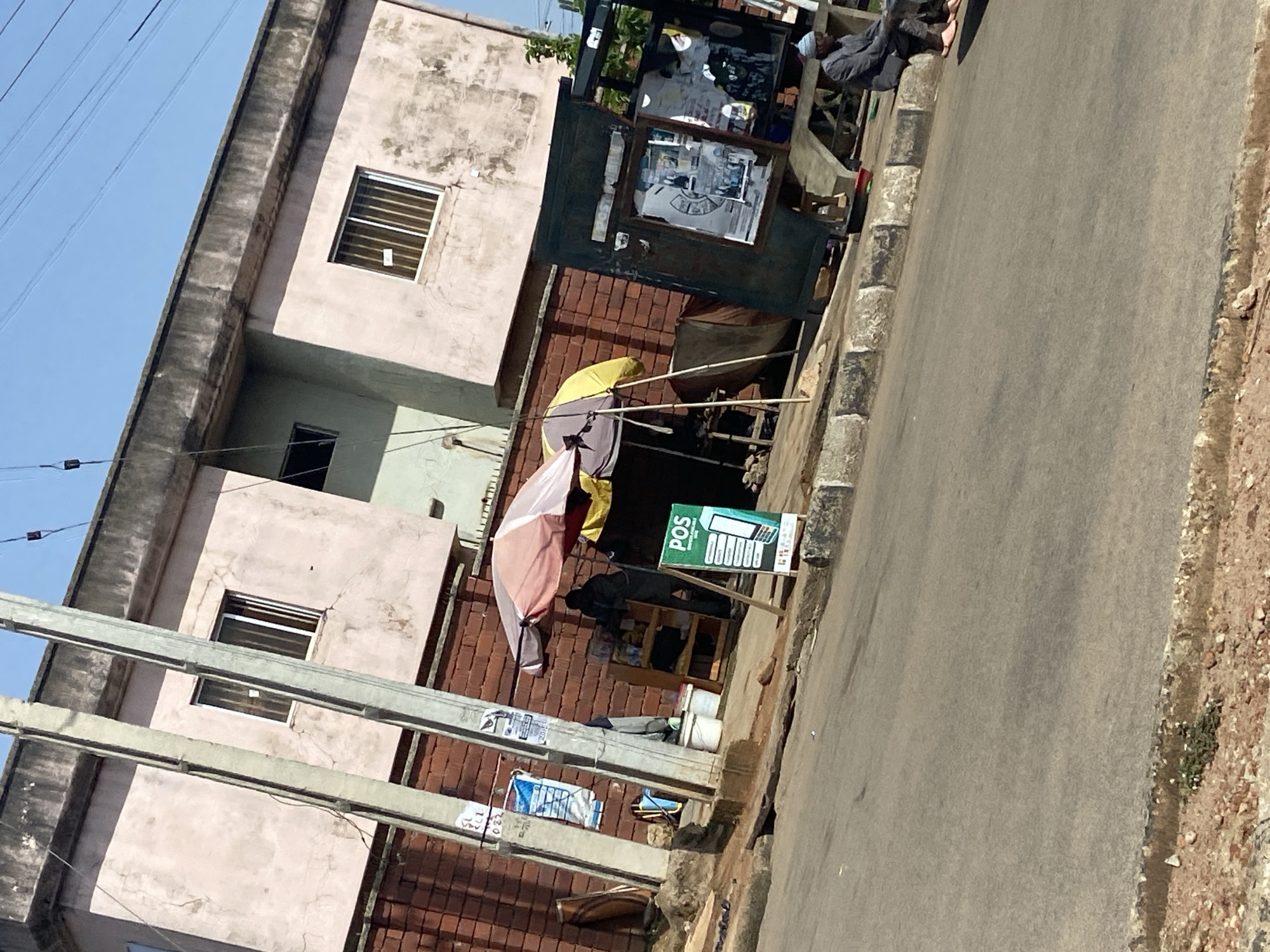 The image depicts a street scene in the city of Jos, Nigeria, characterized by a roadside stall and a building in the background. A pale pink, two-story building with visible signs of wear, such as peeling paint and dirt, forms the backdrop. The building has several windows with barred grills and some are partially obscured by power lines. Adjacent to the building is a brick wall.
In the foreground, a street vendor's stall is set up. The stall is partially covered by umbrellas, one with pink and white sections, and another with yellow. A person, dressed in dark clothing, appears to be attending the stall. In front of the stall, a green sign with "POS" and images of a mobile phone and a card is displayed, indicating a point-of-sale service. Nearby, there are two white buckets and shelves stocked with items. To the right of the stall is a dark, enclosed structure, possibly a more permanent kiosk or a vehicle-like booth, with items displayed inside and on its exterior.
A man in a dark suit and a light-colored cap is seated on the curb on the right side of the image, facing away from the stall and towards the road. He is wearing pink sandals.
The street itself is paved with asphalt and shows signs of wear. The roadside is lined with concrete curbs and some gravelly terrain with sparse vegetation. Power lines are visible crisscrossing the sky, supported by tall concrete poles. The overall impression is of an active urban street environment with a commercial presence.