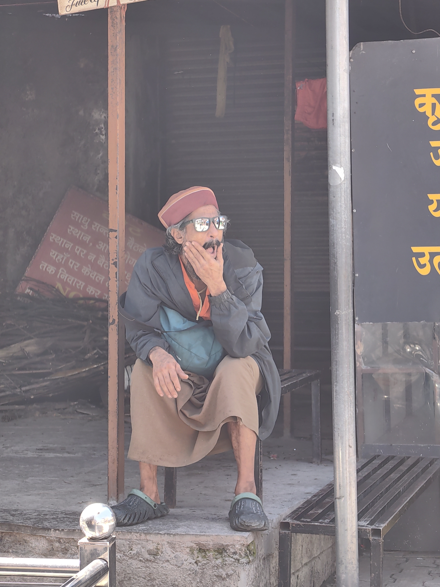 A man is sitting on a bench in Vashisht, India, with his hand covering his mouth as if yawning or speaking. He is wearing a red cap, sunglasses, a dark jacket over an orange shirt, and brown pants. He has a teal bag slung over his shoulder. To his left, a red sign with white text in an Indian language is visible. Behind him, a corrugated metal shutter is partially visible. To his right, a dark sign with yellow lettering is also present. The man is wearing dark green clogs. A metal post and a shiny spherical object are in the foreground. The environment appears to be an outdoor market or storefront.