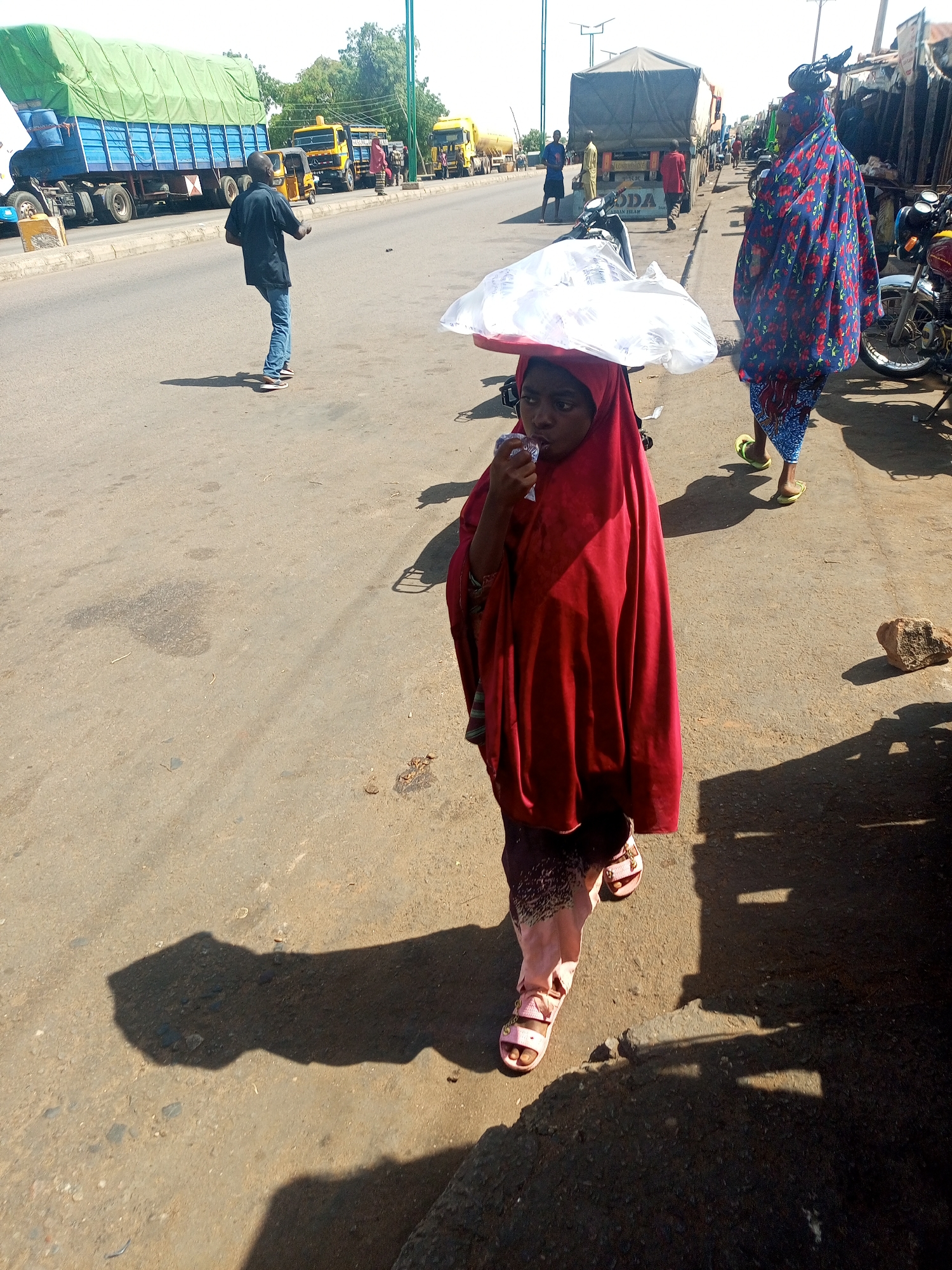The image portrays a bustling street scene in Gombe, Nigeria, captured under bright, clear sunlight, evidenced by the distinct, elongated shadows on the asphalt.
The central subject is a young girl, possibly a child, in the foreground, wearing a red hijab that covers her head and most of her body, over patterned lower garments and pink sandals. She is focused on drinking from a clear plastic sachet while expertly balancing a red tray with white plastic-wrapped items on her head, suggesting she is involved in street vending or carrying goods.
To the right, a woman in a vibrant blue and red patterned full-length dress and lime green flip-flops walks along the roadside, also with an object on her head. Further back on the road, a man in a dark blue shirt and jeans walks away from the camera. The street is a hive of activity, with several large commercial trucks visible in the background, including a prominent blue truck with a green tarp and other yellow trucks. A yellow three-wheeled auto-rickshaw (Keke Napep) is also present. Motorcycles are parked along the right side of the road, adjacent to what appear to be makeshift market stalls.
The scene captures a moment of daily life and commerce, with people moving and goods being transported. On one of the trucks in the background, the text "ODA" and what appears to be "TRANSPORT" are partially visible.
