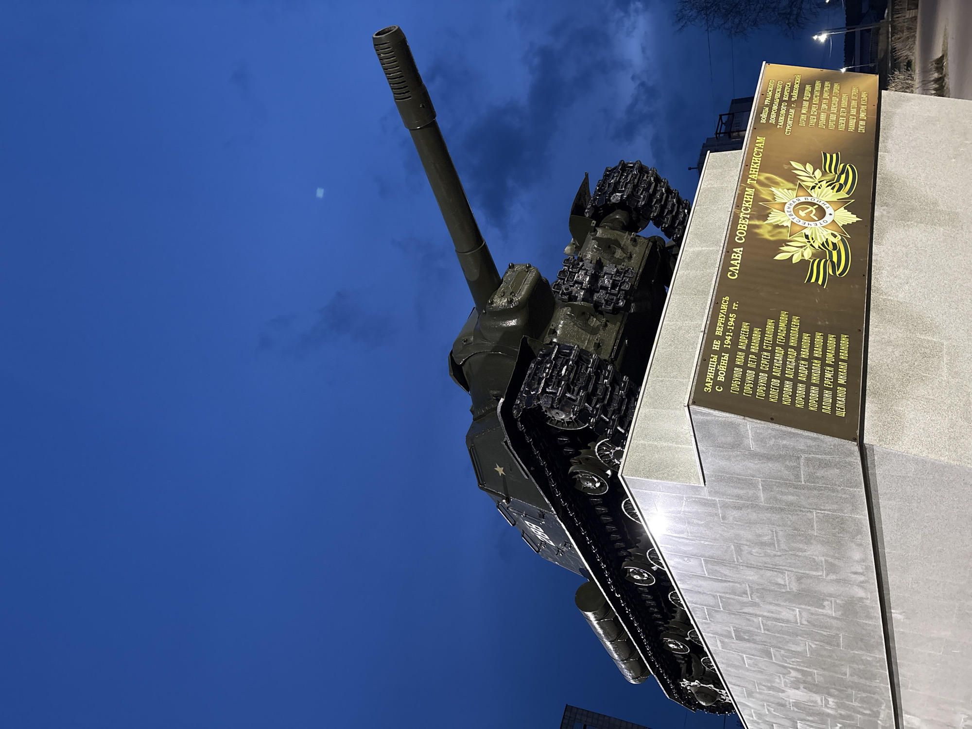 The image shows a Soviet-era tank mounted on a concrete pedestal in Chaikovsky, Russia. The tank is green and faces upwards, with its barrel pointing towards the upper left of the frame. The tank's tracks are visible, as are the wheels along the bottom. A gold-colored plaque is affixed to the right side of the pedestal. The plaque is inscribed in Russian. The text includes the phrase "СЛАВА СОВЕТСКИМ ТАНКИСТАМ" (Glory to Soviet Tankers) and lists names of soldiers who did not return from the war in 1941-1945. The background is a dark blue sky. There is a streetlamp and some trees visible in the upper right corner of the frame.