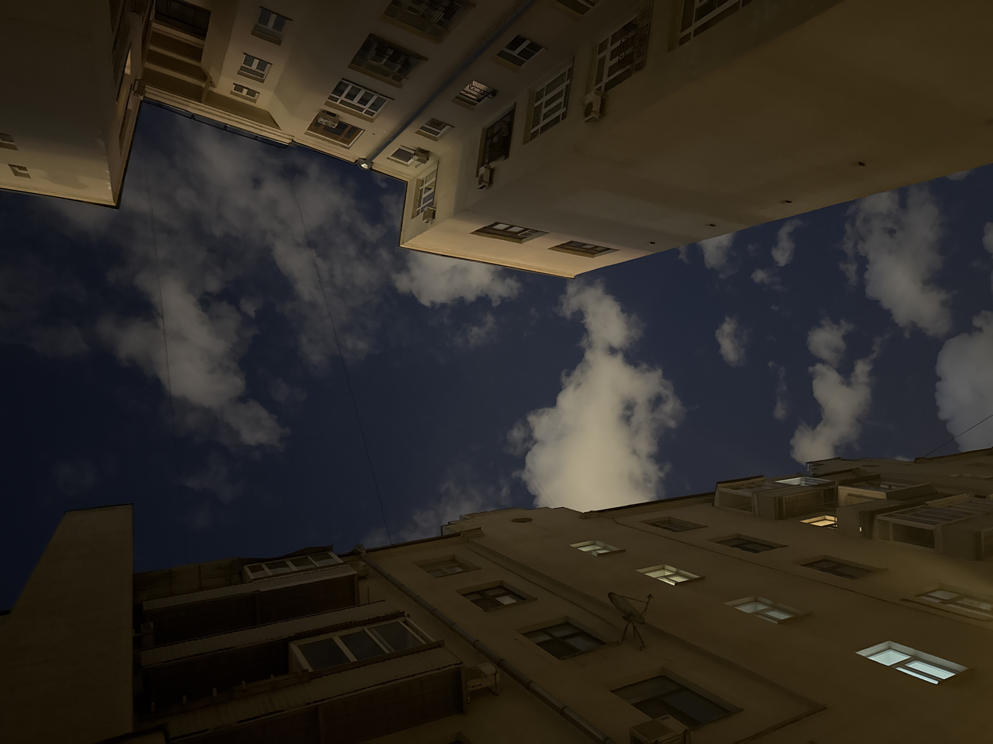 The image presents an upward-looking view into a night sky, framed by the surrounding facades of tall apartment buildings, likely in Moscow, Russia. The sky is a deep, dark blue, adorned with scattered white and grey clouds that are softly illuminated, suggesting moonlight or distant city glow. Thin wires or cables are faintly visible stretching across the sky between the buildings.
Three main buildings are visible, enclosing the sky. The building in the foreground at the bottom right extends upwards, showcasing multiple windows, some of which emit warm, yellow light, indicating occupied rooms. Visible details include balconies, what appear to be drainpipes, and a small satellite dish mounted on the wall. The two buildings in the upper part of the image, one on the top left and another on the top right, frame the upper portion of the sky. These buildings also feature numerous windows and some external air conditioning units. Their facades are a light, warm beige color, illuminated from below, creating a stark contrast with the dark sky.
The perspective is dramatic, giving the impression of standing in a narrow courtyard or shaft, looking directly upwards. There are no people visible, and no specific activity is taking place other than the static scene of the buildings against the night sky. The time of day is clearly night or late evening, as indicated by the dark sky and the artificial lights from windows. No visible text is present in the image.