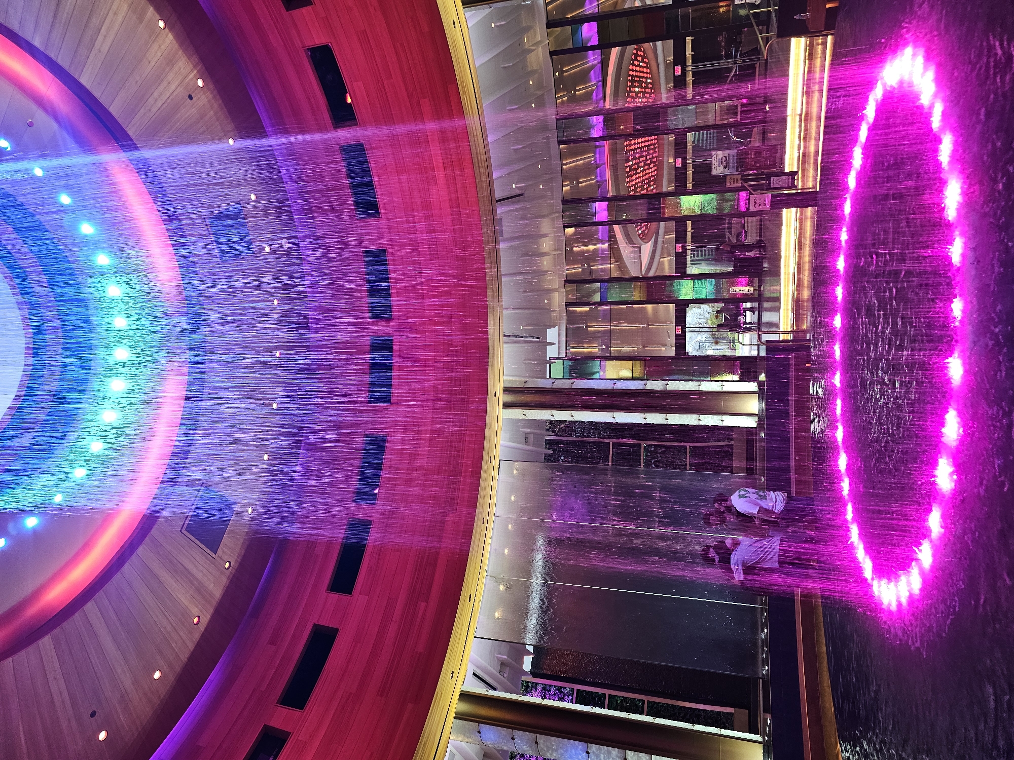 The photograph depicts an interior space within the Seminole Hard Rock Hotel & Casino Hollywood, located in Hollywood Seminole Reservation, United States of America.
A major element is a multi-colored fountain featuring strands of water illuminated in hues ranging from blue to pink and purple. The fountain has a circular design and the cascading water forms a visual screen.
In the foreground, two individuals are standing on a platform. They are positioned near the fountain, appearing to be within its range. Their attire includes white tops. The angle suggests they are likely observing or interacting with the water feature.
The background consists of architectural elements of the interior of the casino and hotel. These include reflective surfaces, a mezzanine with glass barriers, and design motifs typical of the Hard Rock brand. The walls and ceilings incorporate panels in shades of red, brown, and gold. A ring of lights are along the side of the building. The scene is well-lit with colored lights, highlighting the fountain.