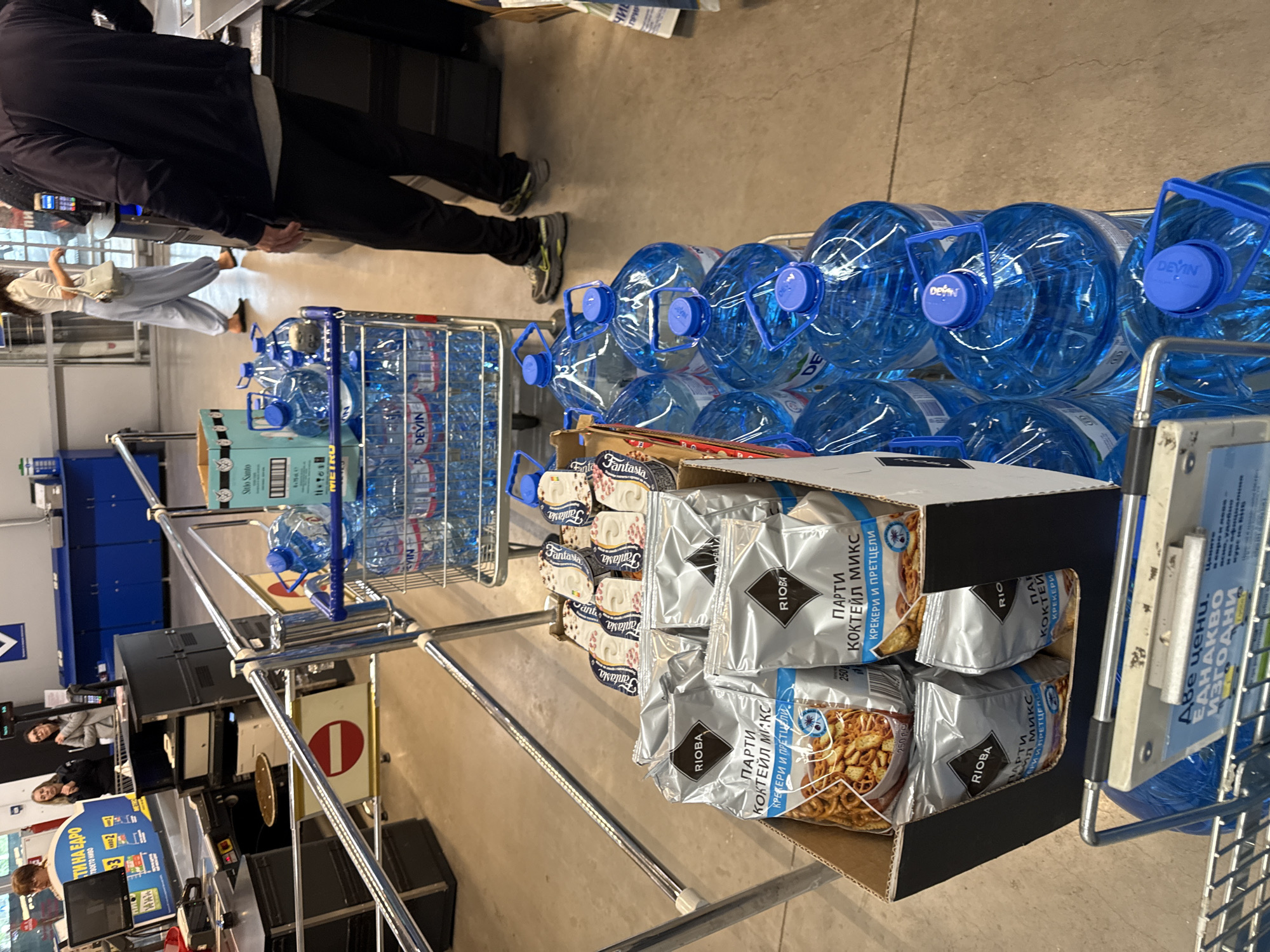 The image shows a shopping cart filled with bottled water and boxes of snack mix. The bottled water is in large blue containers with blue caps. The snack mix is in silver packages labeled "RIOBA" with the words "ПАРТИ КОКТЕЙЛ МИКС КРЕКЕРИ И ПРЕТЦЕЛИ." In addition, there are stacks of "Fantasia" snack mixes. The cart is in a checkout area, with a person standing beside it. In the background, there are people and checkout stands with merchandise on display. In the lower left, there is a sign reading "СТИ НА Е А ЕДРО ТВОЕТО НА 3 овин о". In addition, a sign in Cyrillic has the words "Две цени. ЕДНАКВО ИЗГОДНИ". The scene appears to be in a large retail store, possibly a Metro store, in Burgas, Bulgaria.