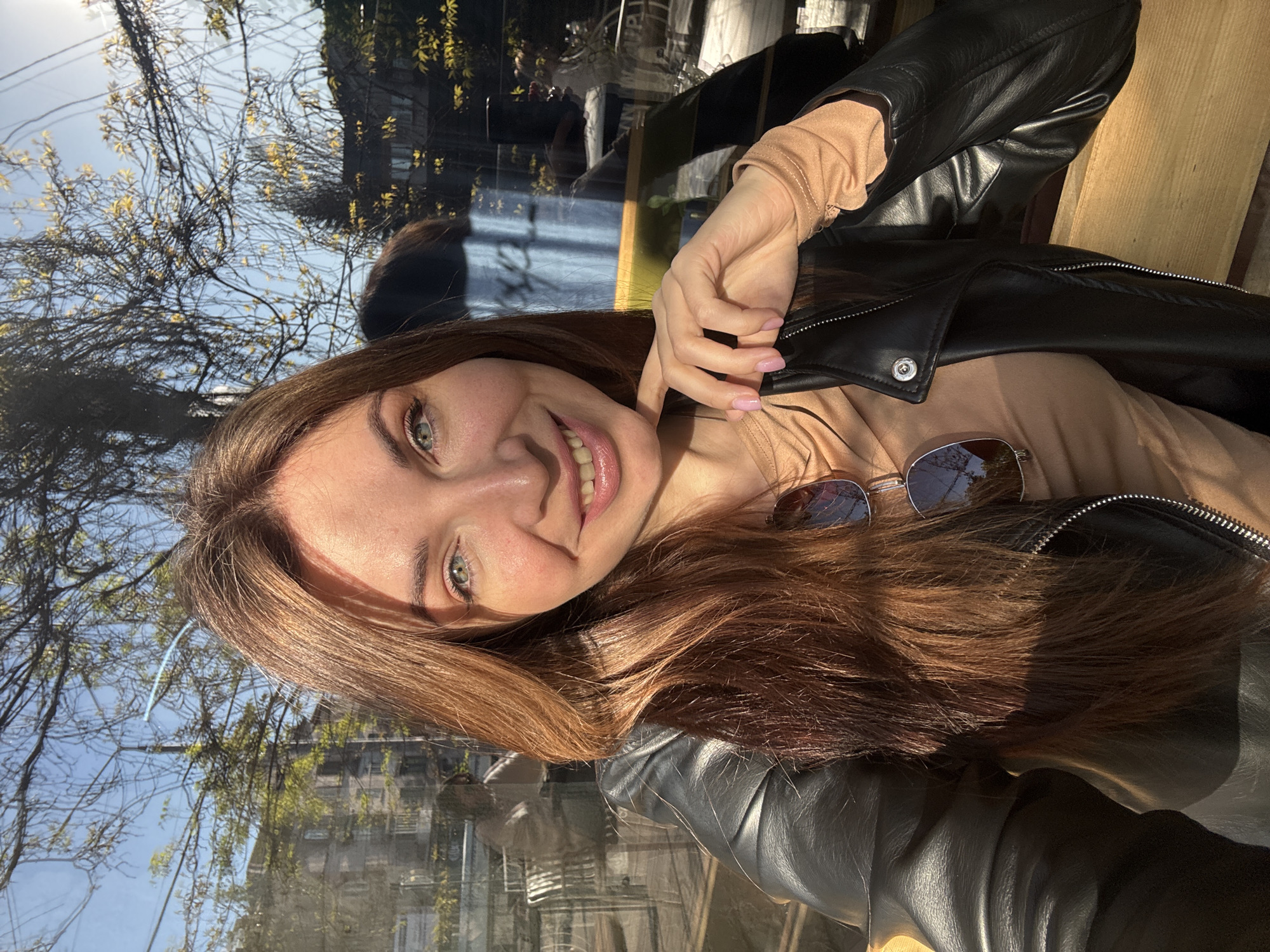 A woman is smiling while seated in what appears to be a restaurant. She is wearing a black leather jacket over a nude-colored top and sunglasses are placed on the top of her clothing near her shoulder. The window behind her reflects the outside environment, including trees and buildings, possibly in an urban setting. While the scene appears to be an urban setting, it is not possible to determine the location in Kryvyi Rih, Ukraine from the image alone.