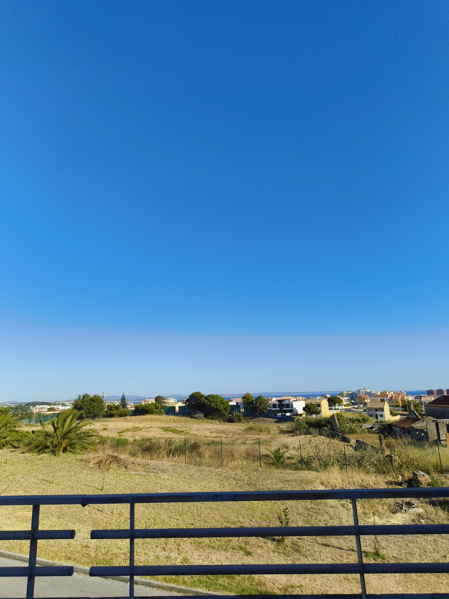 The image shows a daytime, outdoor scene in Cascais, Portugal, looking out over a residential area. In the foreground, there is a dark metal railing. Beyond the railing is a field with dry, light brown grass and some scattered bushes and small trees. There are also some palm trees visible. In the middle ground, there is a collection of houses and buildings with tiled roofs, some appearing to be under construction or in disrepair. The houses are of various colors, including white, yellow, and red. Further in the background, the sea is visible, along with distant structures and the horizon. The sky is a clear, bright blue.
