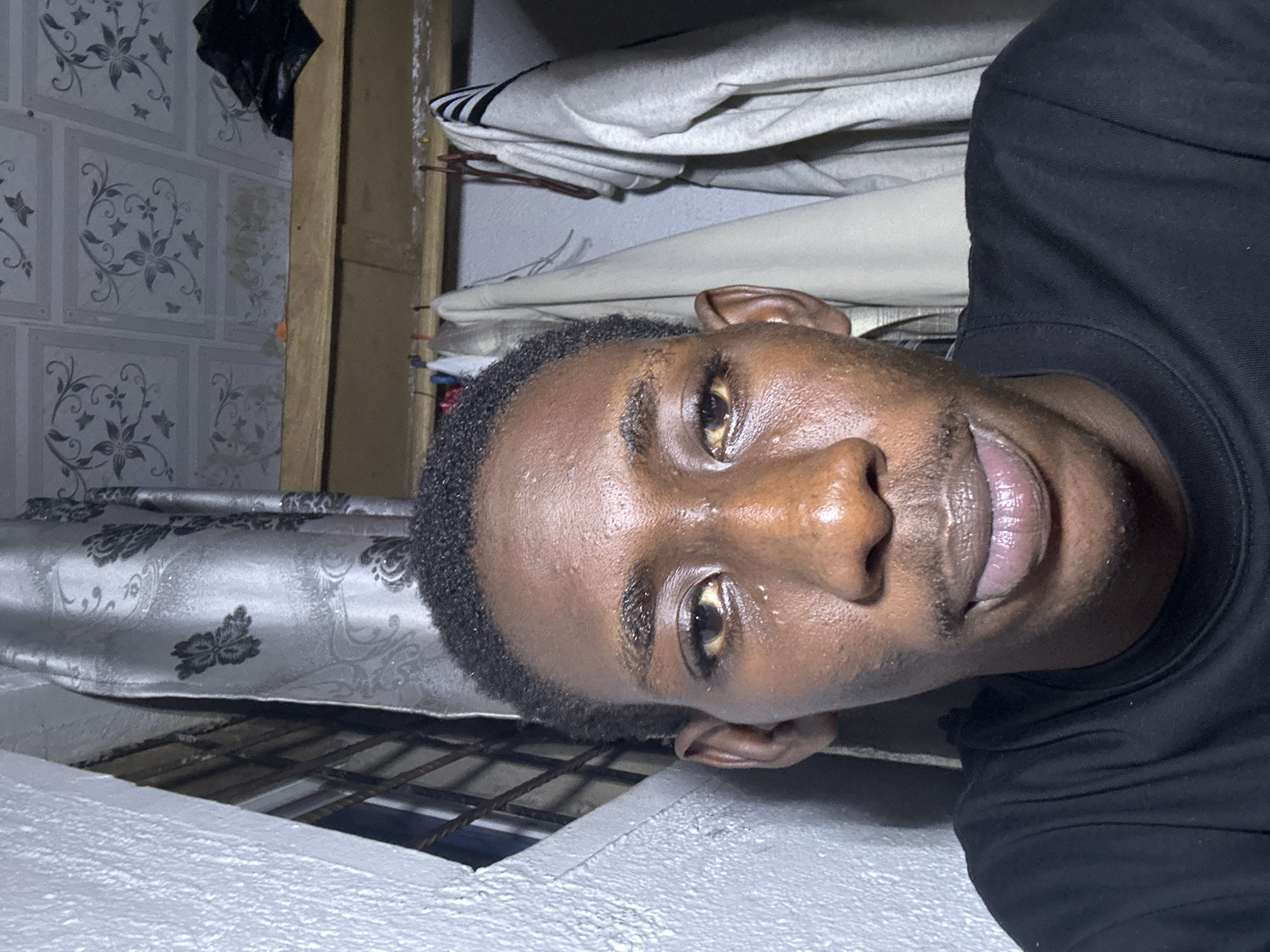 A head-and-shoulders, close-up, selfie-style photograph shows a young Black male with dark skin, short dark hair, and a thin mustache. He is wearing a black t-shirt. His face is close to the camera, and his gaze is directed forward. The background is a dimly lit interior space, featuring a window with bars and a patterned curtain to the left, and a shelf with clothing hanging on it to the right. The ceiling appears to be tiled with a floral pattern. The setting is consistent with a residential interior in Durumi, Nigeria.