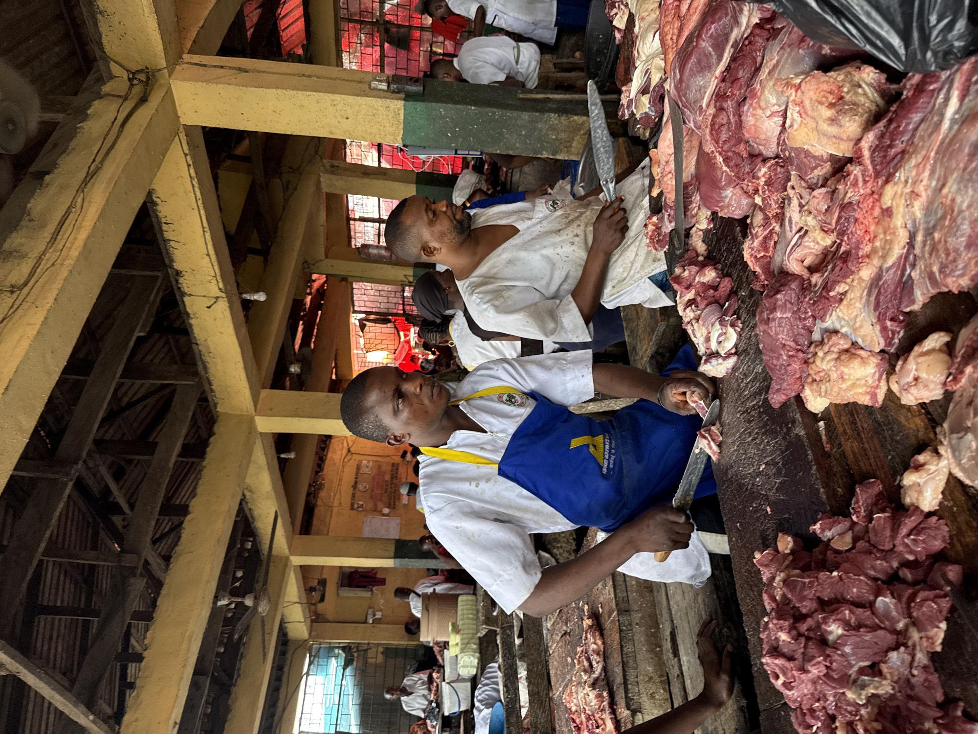 This image depicts a bustling indoor meat market, likely in Lekki, Nigeria, given the context. The primary subjects are several male butchers engaged in processing and selling large quantities of raw meat.
In the foreground, a butcher wearing a white shirt and a blue apron with a yellow logo and text (partially reading "LAGOS WORKMANSHIP...") holds a large knife, appearing to cut or prepare meat on a thick wooden block. Beside him and extending into the mid-ground are substantial piles of fresh red meat, primarily beef, with visible muscle, fat, and connective tissue. Another butcher, in a white shirt, stands behind a similar spread of meat, also holding a large knife. A black plastic bag is seen resting on a pile of meat in the upper right corner.
The market interior features robust construction, with prominent yellow and green-painted concrete beams supporting a network of wooden rafters and a corrugated metal roof. Wires with small light bulbs are strung between the beams, providing illumination. Red-tinted window-like openings or screens are visible in the background, suggesting ventilation or partitions within the market structure. A blurred fan is visible in the upper left. In the background, other market activity and figures are discernible, and a poster on a wall partially displays "LAGOS STATE" text, consistent with the location context. The scene conveys a busy, functional environment dedicated to meat preparation and sales.