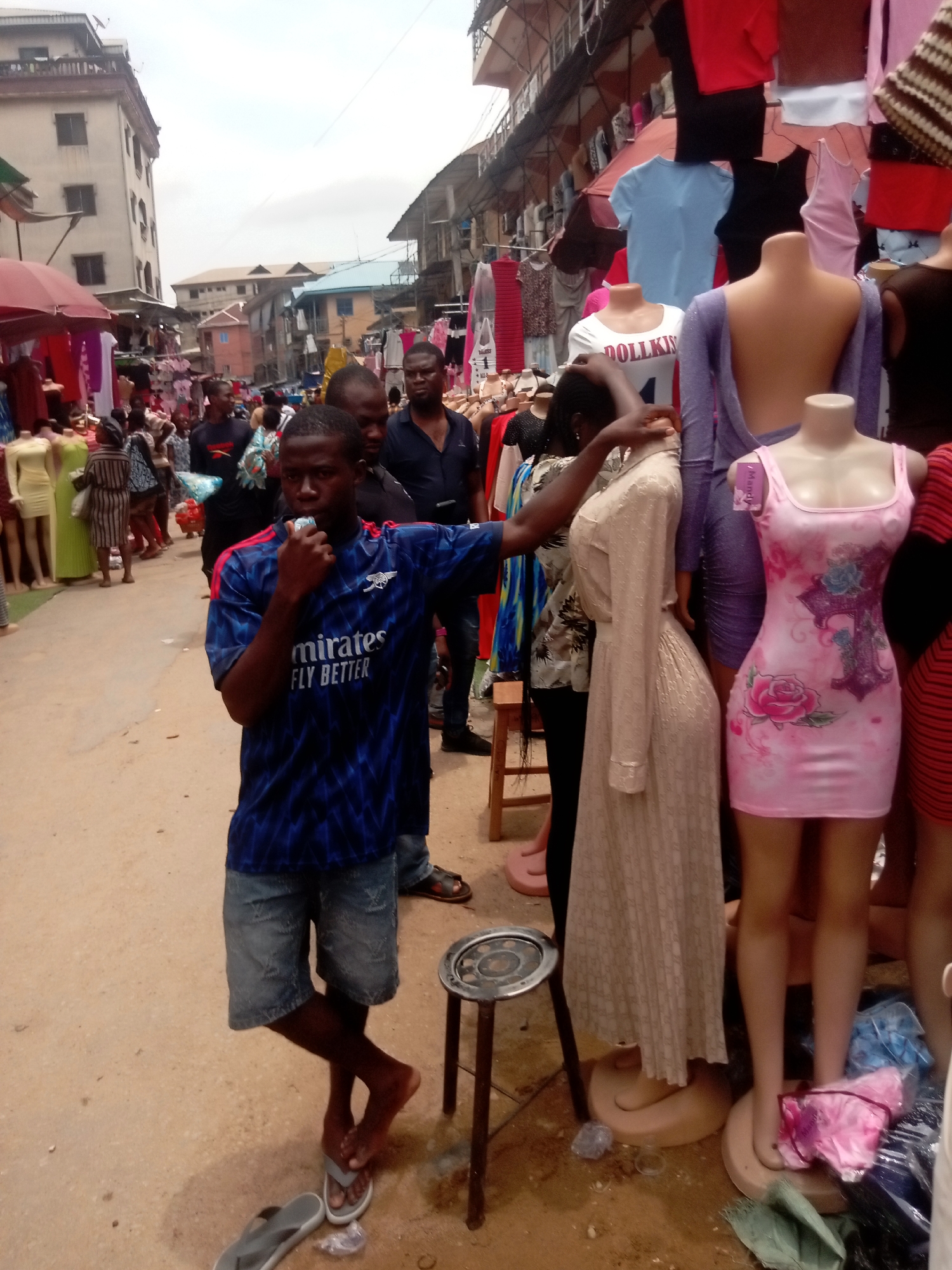 The image captures a busy street scene, likely a market area, in Fegge, Nigeria. In the foreground, a young man wearing a blue Arsenal football jersey and denim shorts stands near a display of women's clothing mannequins. He is holding an object to his mouth, possibly a microphone or whistle, and gesturing with his other hand. Several other people are visible in the background, suggesting a crowded environment. The clothing displayed on the mannequins includes dresses and tops, indicating a retail or market setting. Buildings line the street, hinting at a commercial district. The ground is dusty or sandy, and the overall impression is that of a bustling outdoor market in Fegge.