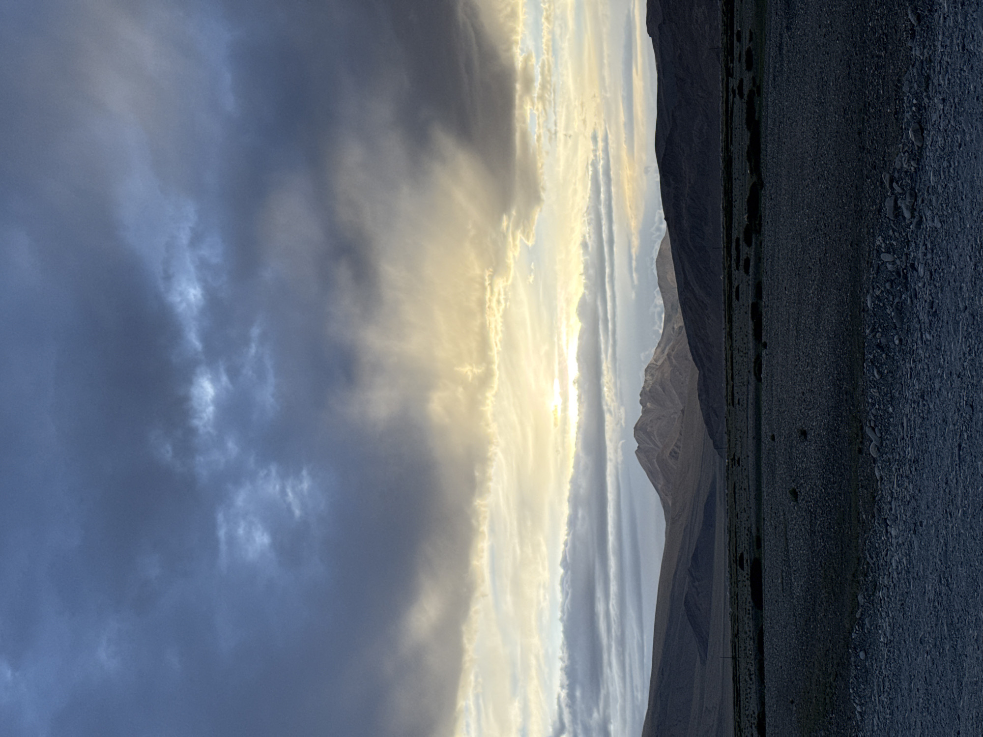 The image depicts a vast, desolate landscape under a dramatic sky. Dark, heavy clouds dominate the upper portion of the frame, with shafts of golden light breaking through them, suggesting a sunrise or sunset. Below the clouds, a range of rugged mountains stretches across the horizon. The foothills are covered in dark, barren terrain, with sparse vegetation visible in the mid-ground. The foreground is a wide expanse of rocky, grey ground, suggesting a dry riverbed or a gravelly plain. The overall impression is one of natural grandeur and isolation, with no signs of human habitation or activity readily apparent. There is no visible indication of the city of Khuldo, India, or any other specific location.