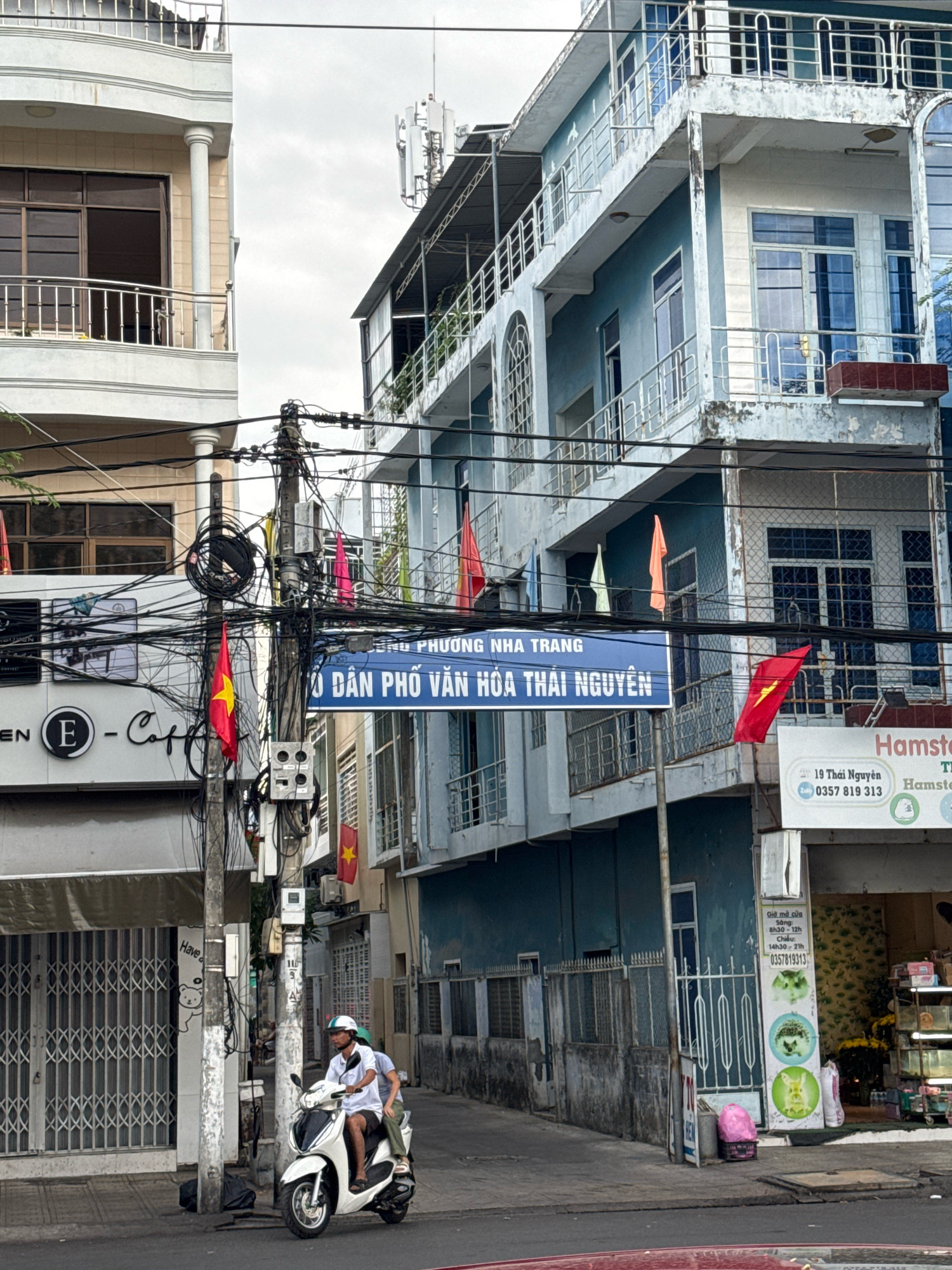 The image captures a street scene in Nha Trang, Vietnam. In the foreground, a white motorcycle with two occupants is moving along the street. The driver is wearing a white shirt and a white helmet, while the passenger is wearing a grey shirt and dark pants. The motorcycle is positioned on the right side of the road.
Dominating the midground are two buildings. The building on the left is a light-colored, multi-story structure with balconies and large windows. The building on the right is a taller, blue multi-story building with balconies and visible windows. A prominent blue street sign is mounted on a utility pole between the two buildings, with text that appears to be in Vietnamese. There are numerous electrical wires and utility poles in front of the buildings. Several red Vietnamese flags are visible, attached to poles and wires.
In the background, more buildings and an overcast sky are visible. The ground is paved, suggesting a street or sidewalk. The environment appears to be an urban setting with commercial establishments visible, including a sign for "Hamster." The overall atmosphere is that of a typical street scene in a Vietnamese city.