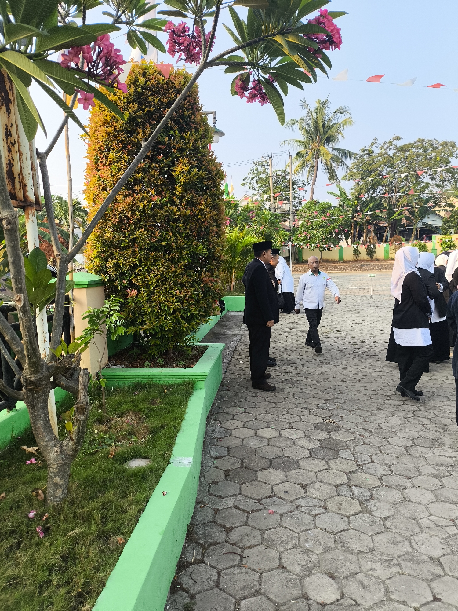 In the city of Cilegon, Indonesia, an outdoor scene unfolds on a hexagonal paved walkway bordered by a vibrant green planter box filled with grass. Several individuals are present, some standing and others walking. On the left, two men in formal dark suits and black caps are positioned near a metal fence and a bare tree trunk with pink flowers blooming above. To their right, a man in a light blue shirt with a green emblem on the chest walks across the pavement, looking to his left. Further to the right, a group of women, some wearing black blazers over white tops and headscarves, stand together. The background reveals lush greenery, trees including a palm, and buildings. Decorative triangular flags in white and orange are strung across the sky. The overall atmosphere suggests a formal gathering or event in a well-maintained outdoor public space.