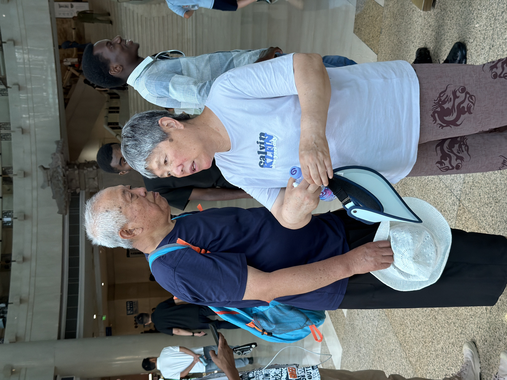 In Yuzhong District, China, two individuals, an elderly man and an elderly woman, are engaged in conversation indoors. The man, with white hair, wears a dark blue t-shirt and black pants and has a blue backpack with orange accents slung over his shoulder. The woman, with gray hair, is dressed in a light purple t-shirt with a "Calvin Klein new york city" logo and patterned brown pants. She holds a white sun hat and a water bottle in her hands. Several other individuals, including two Black men and an Asian man in a white shirt, are visible in the background, along with architectural features such as columns and staircases, suggesting an indoor public space like a museum or shopping mall. The lighting is bright, indicating daytime.