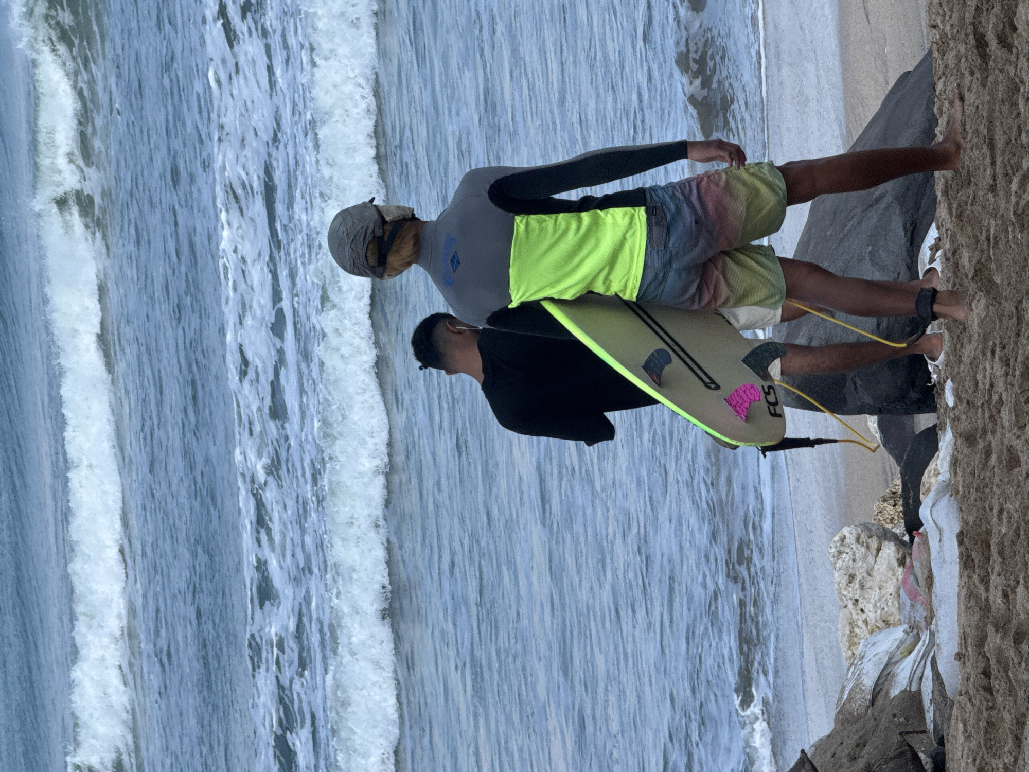 Two individuals stand on a sandy beach, facing the ocean with waves rolling in. One person, on the right, is wearing a long-sleeved rash guard in gray and neon yellow, with multi-colored board shorts and a cap. They are holding a surfboard. The other person, to the left, is wearing a black t-shirt and appears to be holding a phone. The beach has scattered rocks and debris. The scene is the shore of Kuta, Indonesia.