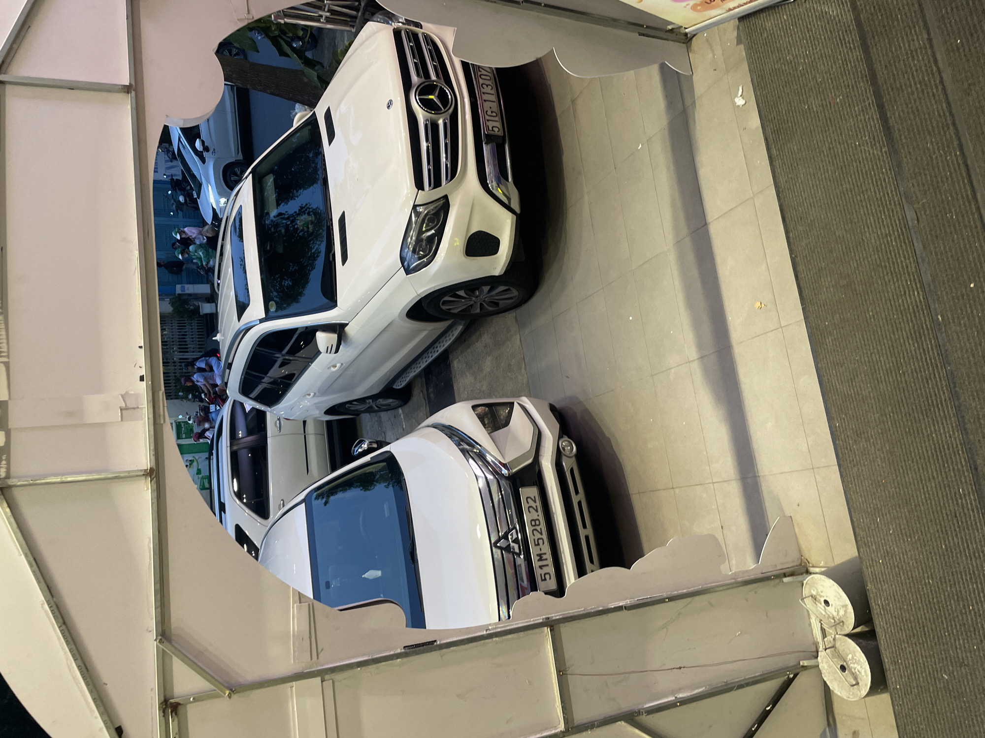 The image captures a street view in Thủ Đức City, Vietnam, focusing on parked vehicles. In the foreground, a white Mitsubishi Xpander with license plate 51M-528.22 is visible. To its right and slightly behind, a white Mercedes-Benz GLS with license plate 51G-11307 is parked. Further in the background, other white vehicles, including a white SUV and a white sedan, are also present. Several individuals are seen on motorcycles on the street, indicating moderate traffic. The environment appears to be an urban setting with paved roads and sidewalks. The scene is viewed from an elevated perspective, possibly from a doorway or balcony, framed by a white structure.