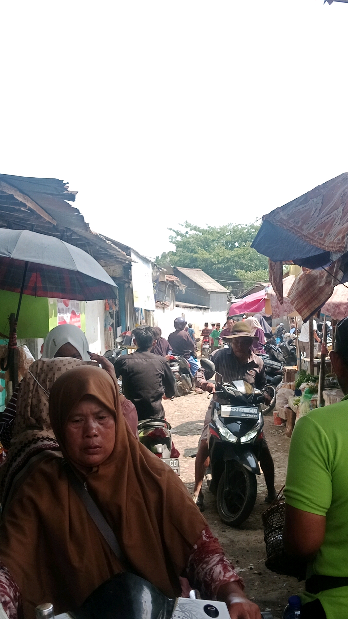 The image depicts a busy street scene, likely a market, in Waru, Indonesia. Several people are present, most of whom are on motorcycles. In the foreground, a woman wearing a brown hijab is seated on a motorcycle. Behind her, another woman in a white hijab is visible. Further back, a man wearing a hat is also on a motorcycle. Other individuals, some of whom are also on motorcycles, are scattered throughout the scene, suggesting activity and movement. The environment appears to be an outdoor market with stalls and temporary structures on either side of the street, indicated by the awnings and shacks. The ground is unpaved, suggesting a rustic or rural setting. The overall atmosphere is one of a bustling local market.