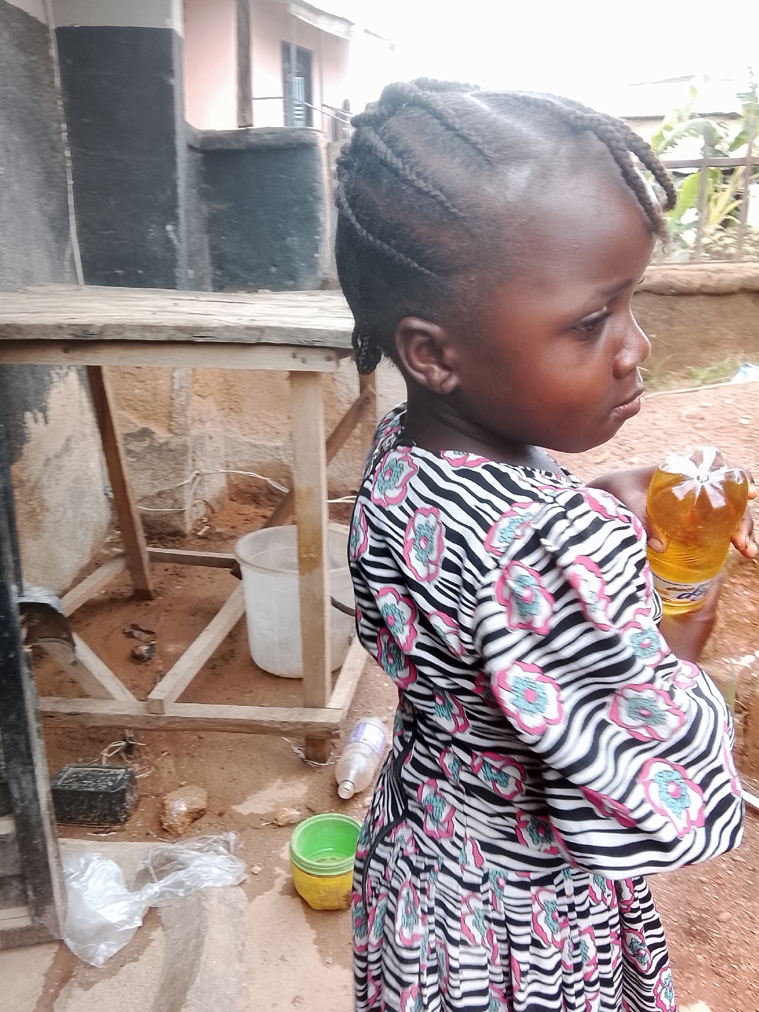 A young Black girl with braided hair stands outdoors in Gwong, Nigeria, holding a plastic bottle filled with yellow liquid. She wears a zebra-striped dress with pink and light blue floral patterns. The scene includes a wooden structure with a bucket underneath, a green and yellow container, and other discarded items on the ground. The background suggests a residential area with buildings and vegetation.
