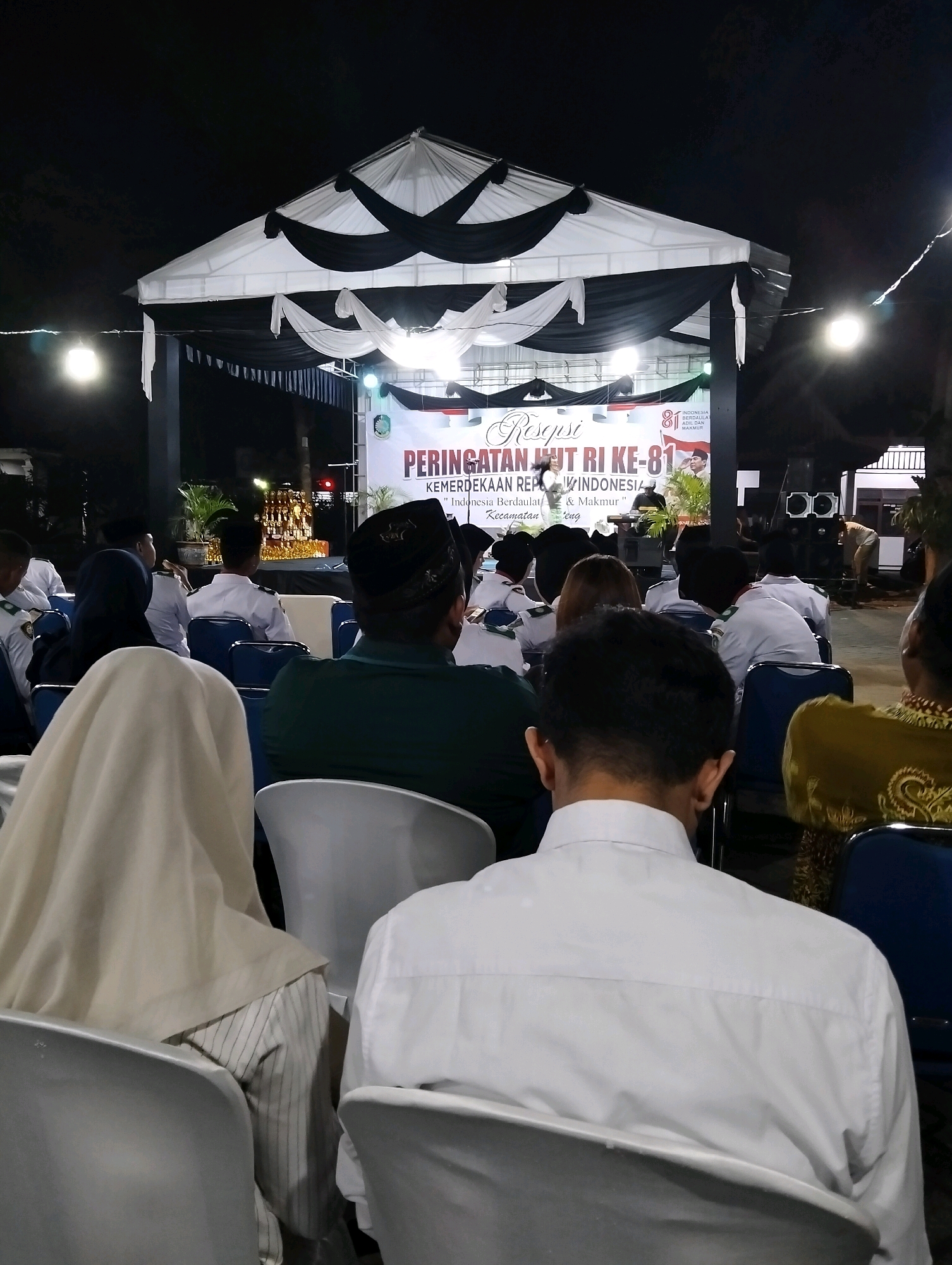 The image shows an outdoor evening event with a stage set up. A white tent with black draping covers the stage, decorated with white fabric streamers. A banner on the stage reads "Resepsi PERINGATAN HUT RI KE-81" and "KEMERDEKAAN REPUBLIK INDONESIA" with smaller text below. There are lights illuminating the stage and the banner. A crowd of people are seated in chairs, facing the stage, with their backs to the camera. Several individuals are wearing white shirts, and some are wearing headscarves or caps. The environment appears to be a public space with some vegetation visible behind the stage. The overall impression is of a formal celebration or ceremony. The location is identified as Gentengwetan, Indonesia.