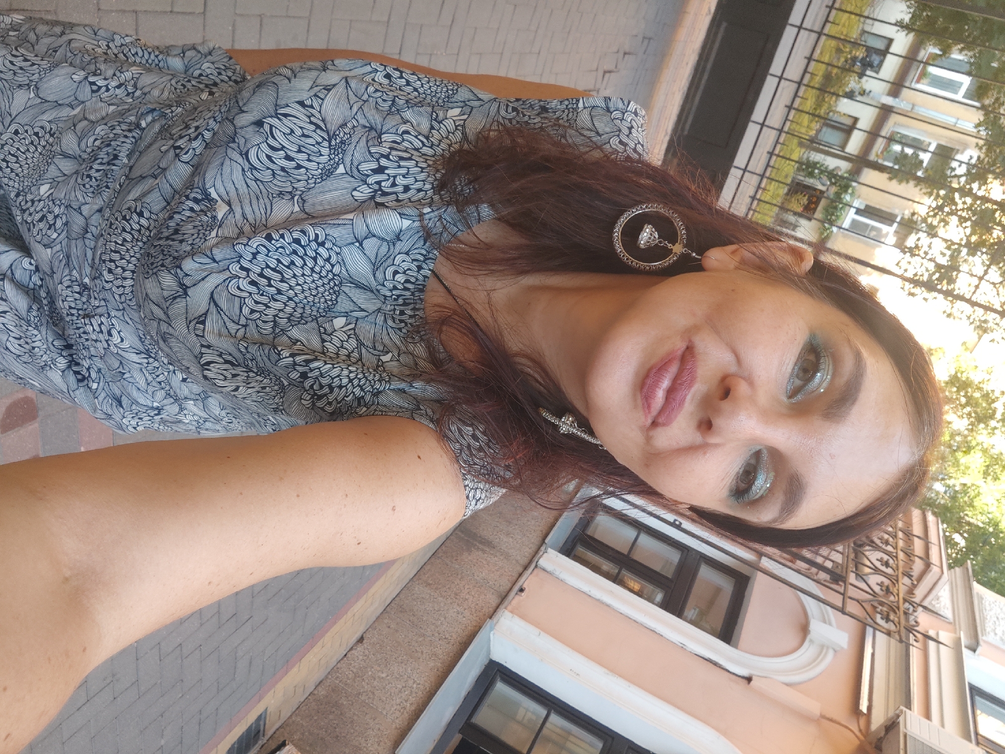 An outdoor, eye-level, medium shot captures a woman with dark hair and prominent blue glitter eyeshadow posing for a selfie. She is wearing a sleeveless blue and white floral print dress and large, circular hoop earrings with a bell-shaped charm. The woman is standing on a paved walkway, with buildings and a black metal fence visible in the background. The lighting suggests it is daytime. The scene is in Saint Petersburg, Russia.