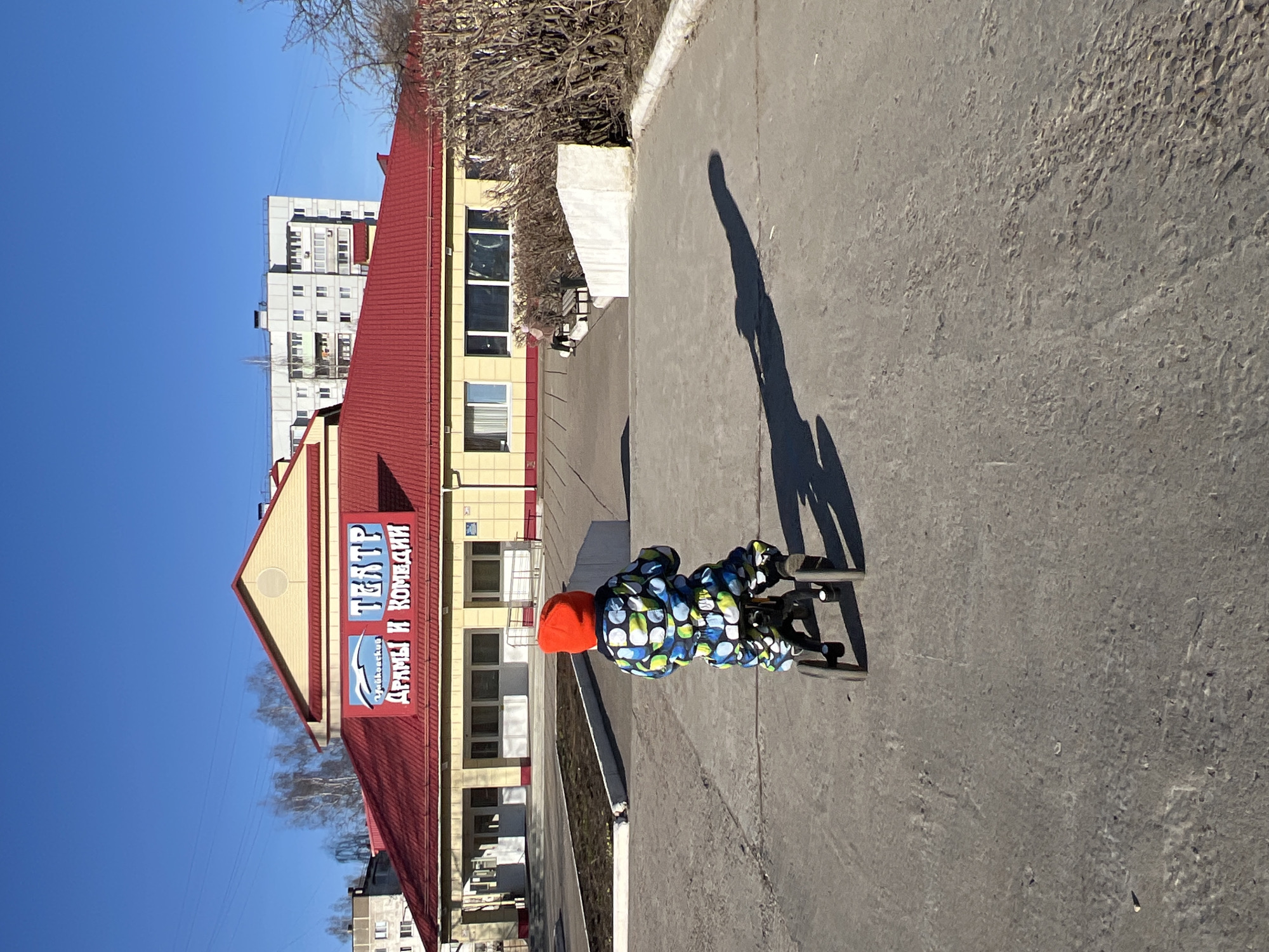 This media file captures an outdoor scene in Chaikovsky, Russia, on a bright, clear day. The primary subject is a young child, viewed from behind, wearing a brightly patterned jacket (blue, green, and white circles) and a vivid orange hat. The child is pushing a black scooter with small wheels along a textured, paved surface, likely asphalt or concrete.
Strong sunlight creates a long, distinct shadow of the child and scooter stretching across the ground. In the midground, a building with a prominent red pitched roof and yellow-beige walls stands out. A large blue and white sign is affixed to the roof, clearly displaying the text "ЧАЙКОВСКИЙ ТЕАТР ДРАМЫ И КОМЕДИИ" (Chaikovsky Theater of Drama and Comedy), with a circular emblem underneath the word "Чайковский". Bare-branched trees or bushes are visible behind a low concrete barrier near the building. In the background, a tall, light-colored apartment building with numerous windows rises against a cloudless, bright blue sky. The scene suggests a pleasant, sunny day, possibly in late winter or early spring, given the bare tree branches.