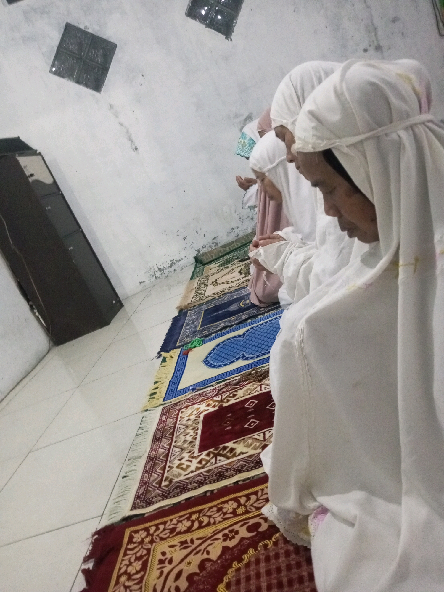 The image depicts four individuals, appearing to be women, in a state of prayer. They are all wearing white head coverings and attire, typical of Islamic prayer garments. They are positioned on prayer rugs laid out on a tiled floor. The setting appears to be an indoor space with plain white walls, some of which show signs of wear or discoloration. A dark, tall cabinet or furniture piece is visible to the left of the frame, and a square, textured glass block is mounted on the wall in the upper left corner. The prayer rugs themselves are colorful and patterned. The overall lighting is dim, suggesting an interior shot, possibly taken at dusk or in a poorly lit room. The image has a candid, observational feel. There is no specific indication of the location being in Andongsili, Indonesia, beyond the prompt's instruction to describe it as such.