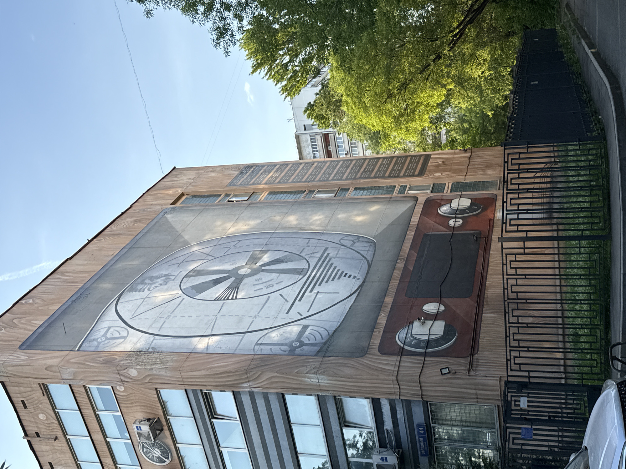Here is a detailed description of the image:
The image shows a building with a mural painted on its side in Moscow, Russia. The mural is designed to resemble an old-fashioned television set. The screen of the "television" displays a test pattern, while the side panel features knobs and dials. The building itself appears to be a commercial or office space, with multiple windows and air conditioning units visible. A blue street sign with white lettering reading "ЗВЕЗДНЫЙ 44" is mounted on the wall of the building; this confirms the street location as Zvezdny Boulevard, building number 44, in Moscow. The surrounding environment includes trees, a fence, and another building in the background. The sky is clear and blue.