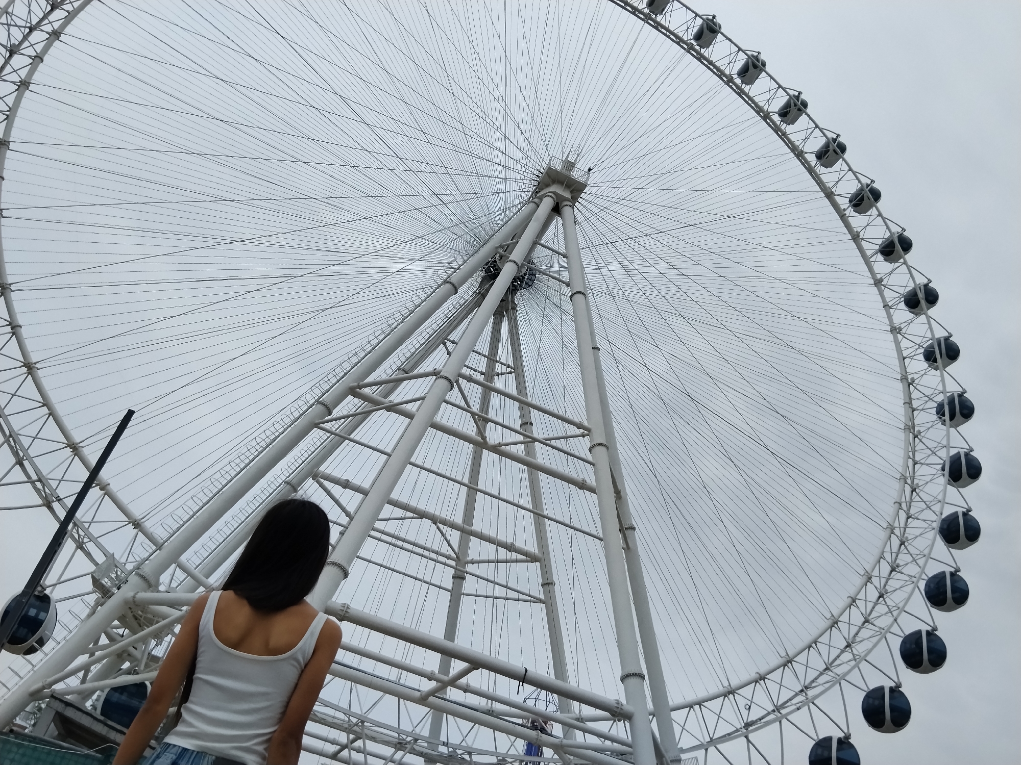 An outdoor, low-angle shot captures a woman standing with her back to the camera, looking up at the complex structure of a large white Ferris wheel against a cloudy sky. The woman has dark hair and is wearing a white tank top and blue jeans. The Ferris wheel dominates the frame, its intricate network of white spokes and cables stretching across the sky. Several dark gondolas are visible attached to the outer rim. The base of the Ferris wheel, with its sturdy white support beams, is also visible in the lower portion of the image. The overall scene suggests a tourist attraction in a city, though the specific location in Bishkek City, Kyrgyzstan is not identifiable from visual cues alone.
