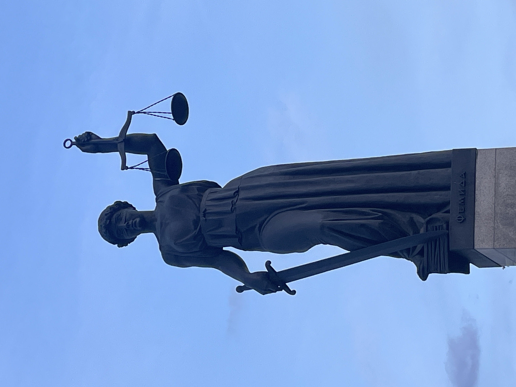 The image shows a statue of Themis, the Greek goddess of justice, located in Krasnoyarsk, Russia. She is depicted holding scales in her raised left hand and a sword in her right hand. The statue is mounted on a pedestal with inscriptions in Russian. The background is a light blue sky.