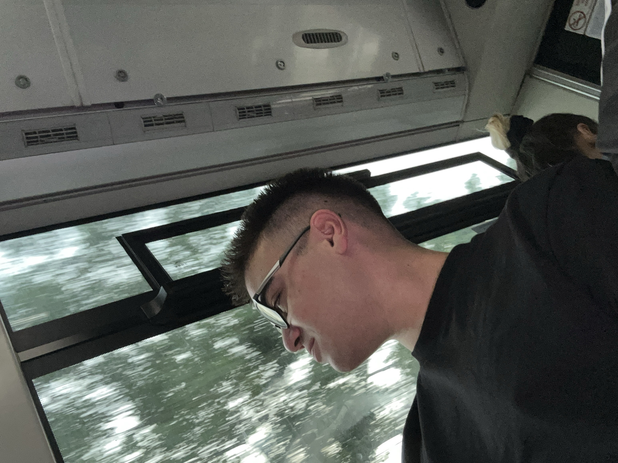 An upward-angled, medium shot shows a young Caucasian male with dark, buzzed hair and glasses looking out a window while riding in a vehicle. The male is wearing a black t-shirt. The window shows a blurred green and white background of trees and sky, suggesting the vehicle is moving. The interior of the vehicle is visible, with white walls and ventilation grates. A second person's head, with a blonde bun, is visible in the lower right portion of the image. The overall lighting is overcast.