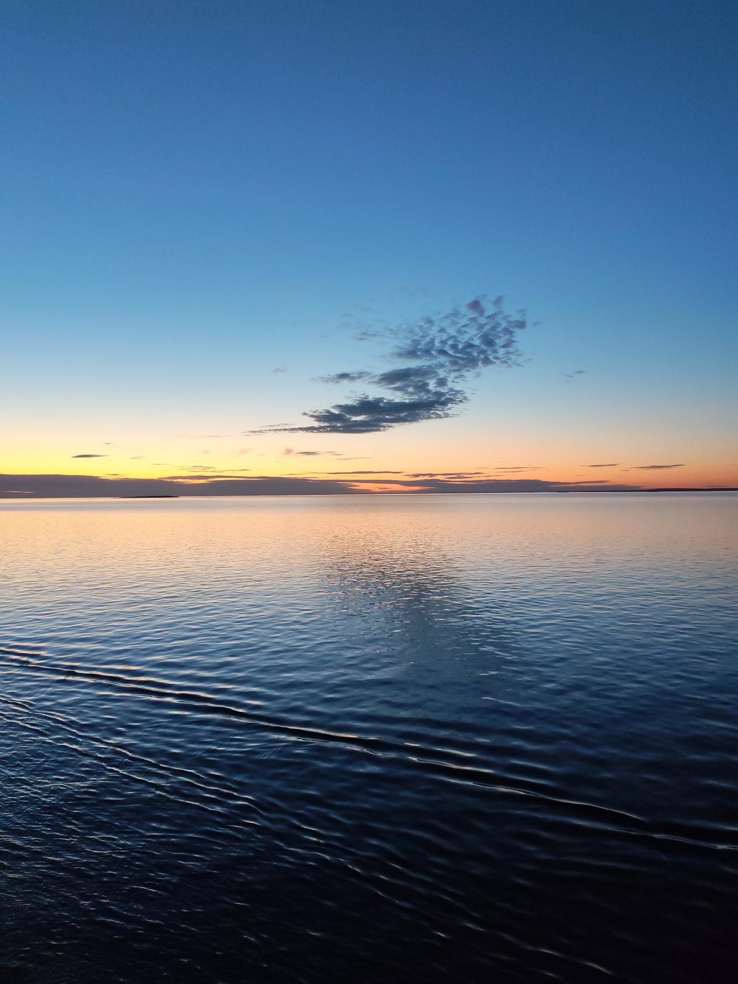 The image depicts a serene seascape or large lake at either dawn or dusk. The sky dominates the upper portion, transitioning from a deep, rich blue at the zenith to lighter blues, then warm yellows and oranges closer to the horizon, indicative of the sun's low position. A distinct cluster of small, textured clouds is visible in the upper right quadrant of the sky, while thinner, elongated cloud formations stretch across the horizon.
The expansive body of water occupies the lower two-thirds of the frame. It reflects the colors of the sky, showcasing warm, golden and orange tones near the horizon, gradually shifting to deeper blues and darker shades in the foreground. The water's surface is not perfectly still; it displays numerous gentle, parallel ripples and small waves, particularly visible in the mid-ground and foreground, creating a sense of subtle movement. A faint, dark landmass or island can be seen as a thin line on the distant horizon.
There are no people, man-made objects, or visible text in the scene. The overall impression is one of tranquility and vastness, capturing a peaceful moment in nature. The weather appears calm with scattered clouds. The location is consistent with a large body of water in "Unknown, Russia."