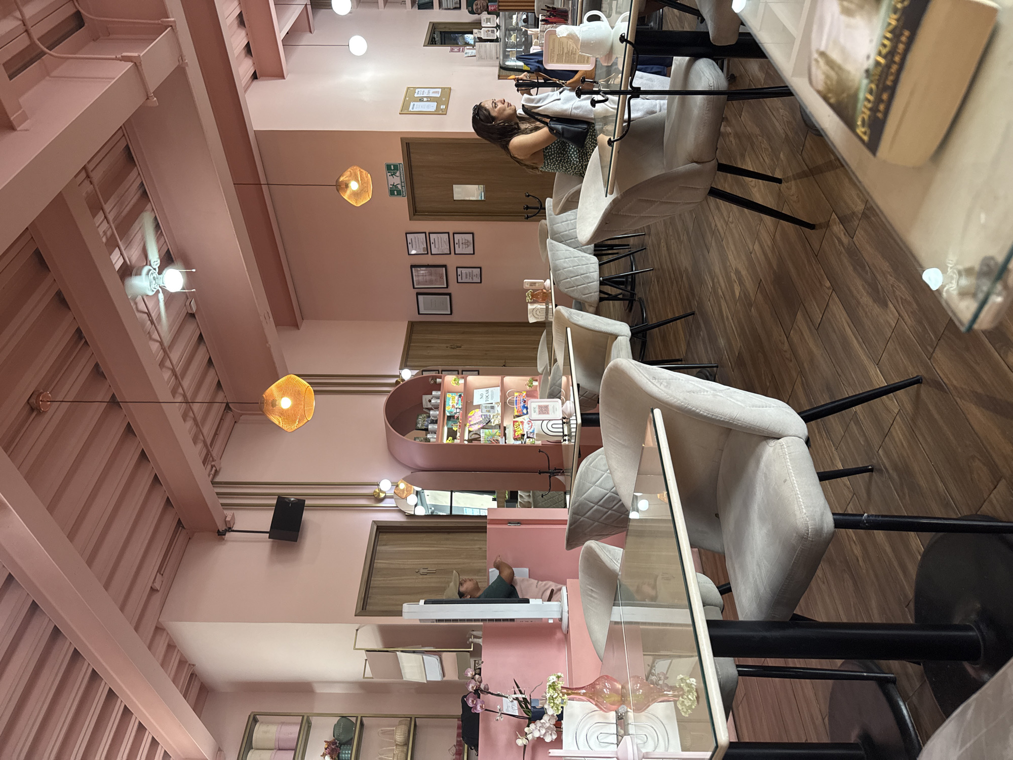 The image depicts the interior of a café or salon located in Puebla, Mexico. The space is decorated in a predominantly pink color scheme with exposed wooden beams on the ceiling. Multiple tables with glass tops and upholstered chairs are arranged in rows. A woman with dark hair wearing a green dress is seated at one of the tables, holding glasses. In the background, there's a shelf with various items displayed. The floor is made of wood planks. Hanging light fixtures with woven shades illuminate the scene. Certificates and other framed items are mounted on the wall. The objects on the tables include decorative items and a book with the title "Lord of the Rings".