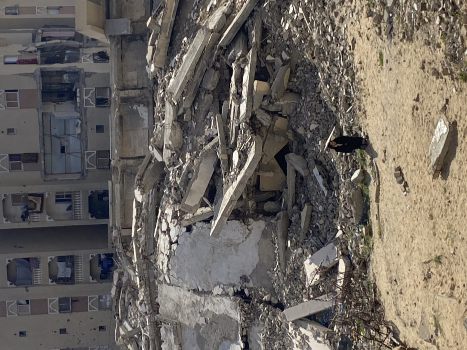 This image depicts a scene of extensive urban destruction in Khan Yunis, Palestinian Territory. The foreground consists of a sandy, light-brown patch of ground with sparse vegetation and scattered small debris. In the midground, a substantial, irregular mound of gray and off-white concrete rubble, rebar, and shattered building materials dominates the view, marking the site of a collapsed or demolished structure.
In the background, behind the rubble pile, stand multiple multi-story residential buildings, beige or light-brown in color, exhibiting significant damage. Several sections of the buildings' facades are missing, exposing internal rooms and structural elements. Bullet holes are visible on the exterior walls of some buildings. Balconies are present, with items resembling laundry or tarps hanging from some.
One individual is visible in the lower-right portion of the image, positioned on the sandy ground. The person is dressed in dark clothing and holds a light-colored object, possibly an umbrella or traditional head covering, over their head, casting a distinct shadow. The person appears to be stationary, either observing the scene or resting. The environment is brightly illuminated by natural sunlight, indicating daytime conditions.