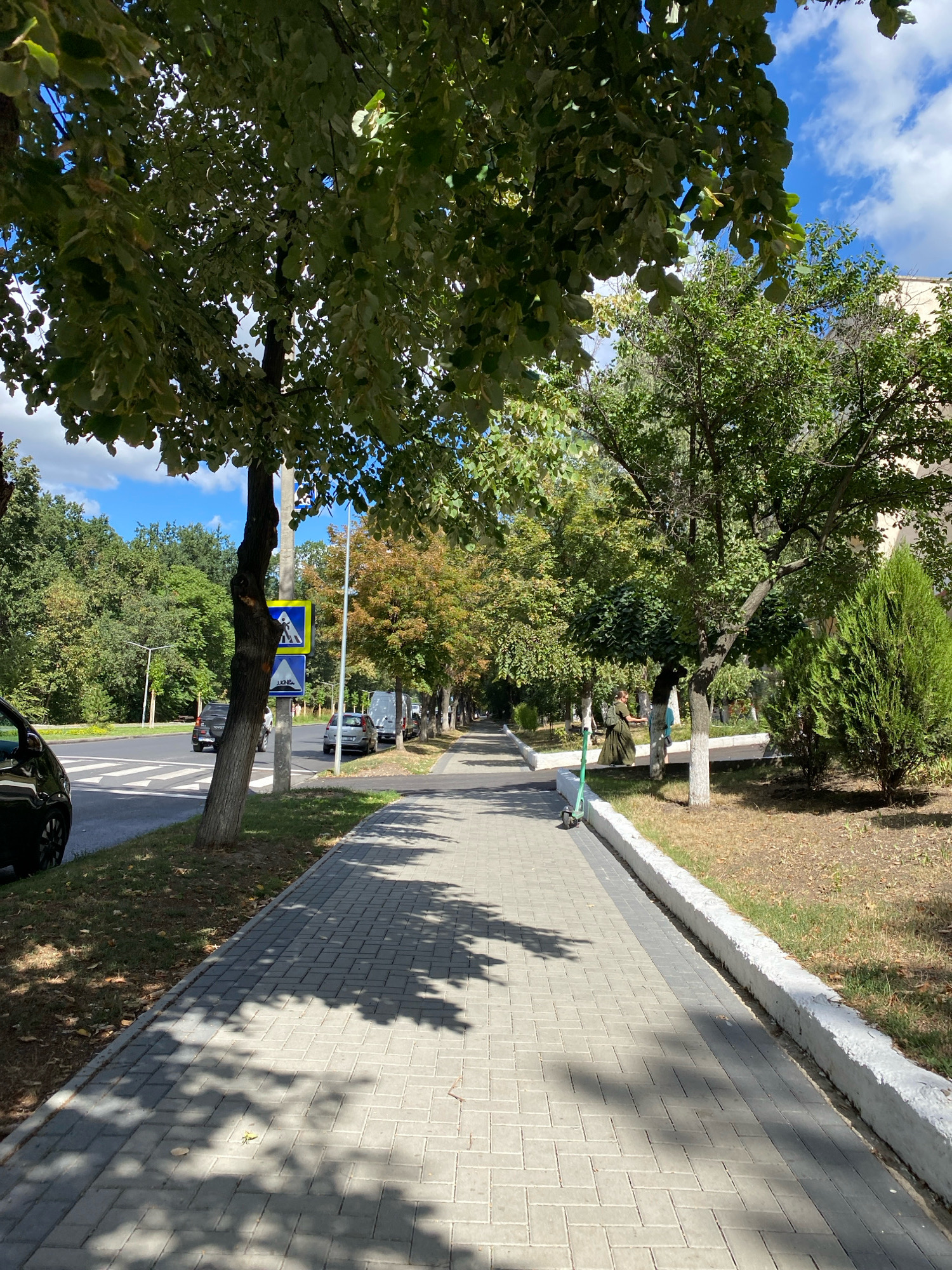 The image depicts a tree-lined sidewalk adjacent to a road with pedestrian crossing markings. The sidewalk is paved with light grey bricks and features a white border. Shadows from the trees are cast across the sidewalk.
On the sidewalk, a green electric scooter is parked. Further down, a person in a dark green dress with a backpack is walking, holding what appears to be a smartphone. Another person is partially visible further down the sidewalk.
The road shows pedestrian crossing markings and traffic lanes. Several cars are parked or driving on the road, including a black car on the left and a silver minivan and a silver car further down. There are traffic signs visible, including a blue and yellow sign with a pedestrian symbol and another sign with a slope icon.
The environment is characterized by lush green trees lining both sides of the road and sidewalk. The sky is blue with scattered white clouds. The overall setting appears to be an urban or suburban street in Chișinău, Moldova, given the context provided. The area seems to be well-maintained, with trees providing shade and a clean sidewalk.