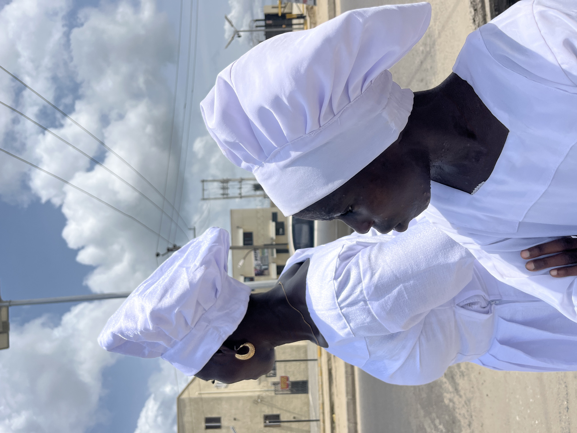 Two people of African descent stand outdoors, wearing white robes and chef's hats. The first person is in the foreground, viewed from the shoulders up, looking down. The second person is in the background, similarly dressed and facing the same direction. A city street with buildings and utility poles is behind them. The sky is partly cloudy. The location appears to be in the city of Ado, Nigeria, as per the prompt.