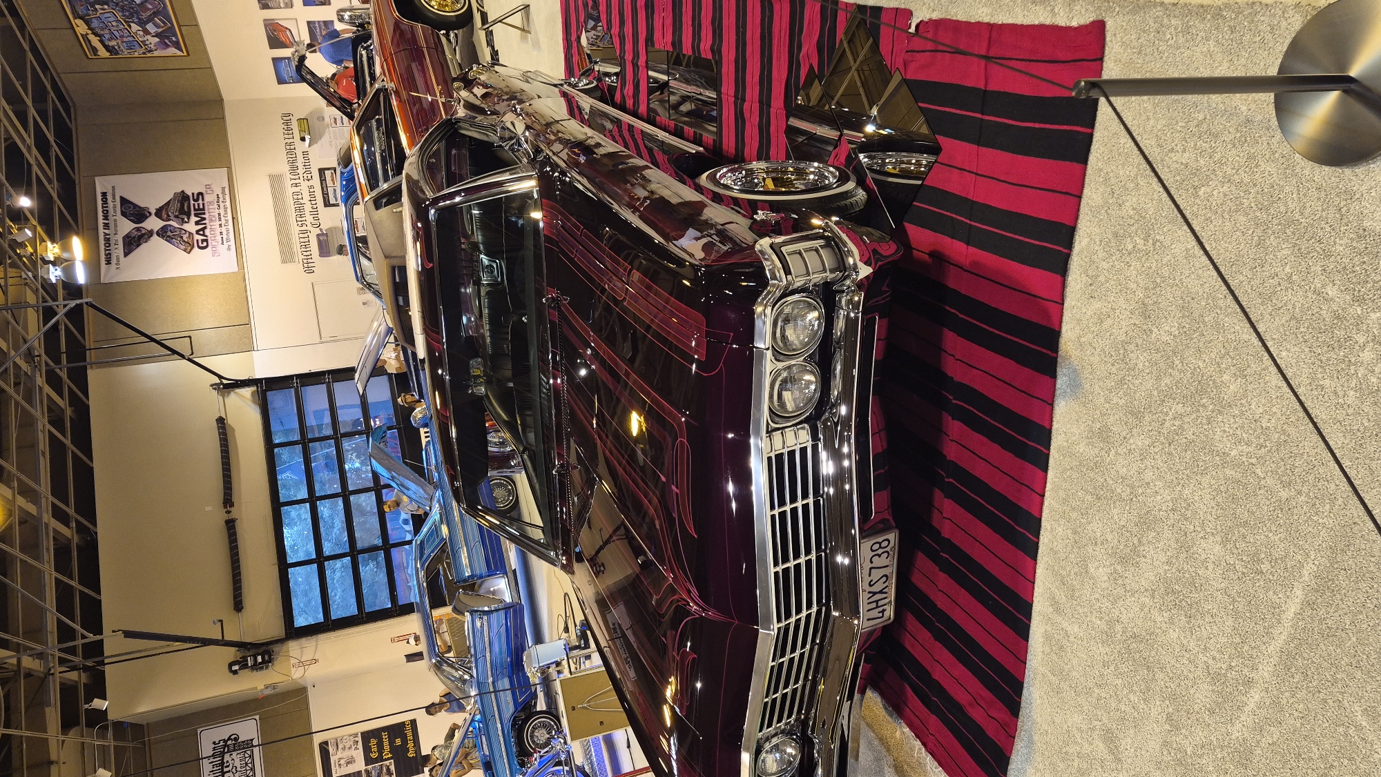 This image captures a lowrider car show, likely held indoors, with a dark purple, classic American car prominently displayed in the foreground. The car features intricate pinstripe detailing in a lighter red hue on its hood and roof, and is positioned on a striped rug with alternating red and black bands. The car's license plate reads "4HXS738". In the background, other lowrider cars are visible, including a blue one with its hood open and a reddish-orange one. Several people are present, observing the vehicles. Above the cars, banners are displayed on the wall; one banner reads "HISTORY IN MOTION" with "GAMES SACRAMENTO, CA" and dates below it, indicating the event took place in Sacramento, California. The environment appears to be a large indoor space with high ceilings, industrial lighting, and large garage-style doors in the background.