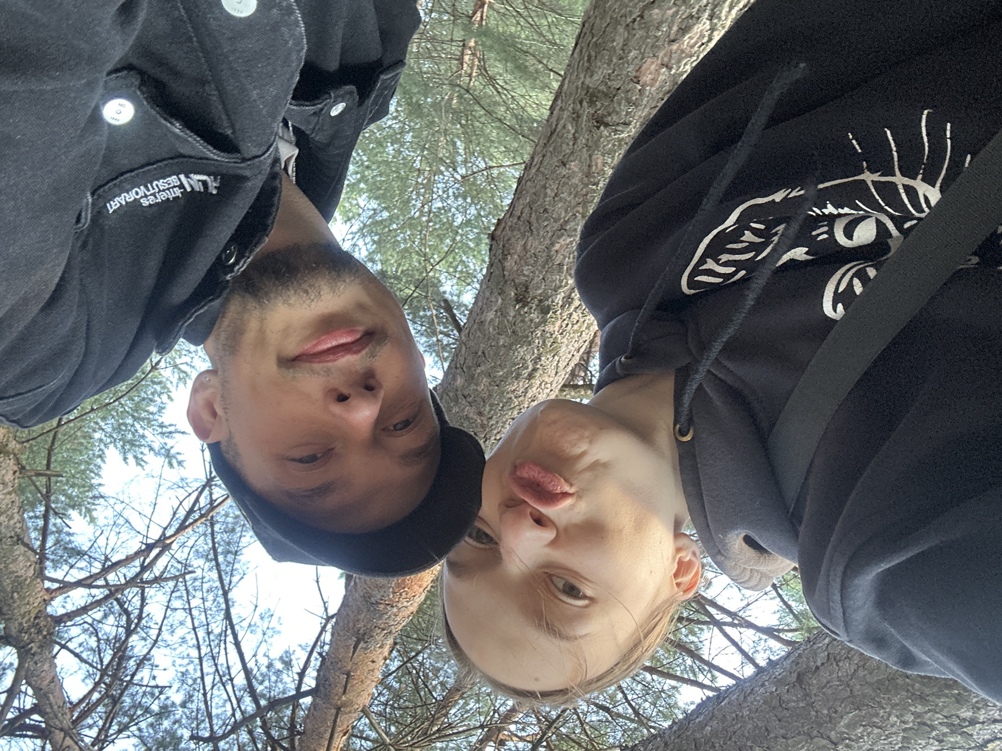 This media file is a selfie taken from a low angle, looking up towards two people among trees and branches. The photo is oriented vertically with the subjects upside down relative to a standard orientation.
The subjects are a man on the left and a woman on the right. The man is wearing a black jacket, possibly denim, with some visible buttons and a collar. He has a beard and is looking towards the camera. On his jacket, the text "RESTUVORART" and "MMC O 1989" are visible. He is also wearing a black cap.
The woman on the right is wearing a black hoodie with a white graphic design on the chest, possibly a stylized animal face. She has blonde hair and is making a puckered expression towards the camera, a common "duck face" pose.
The setting appears to be outdoors, amidst pine trees, with branches and needles visible in the background against a bright sky. The lighting suggests it is daytime, likely sunny. The overall mood is casual and playful, indicated by the selfie pose and the woman's expression. The location context of Chaikovsky, Russia, does not provide any visually discernible cues within the image itself.