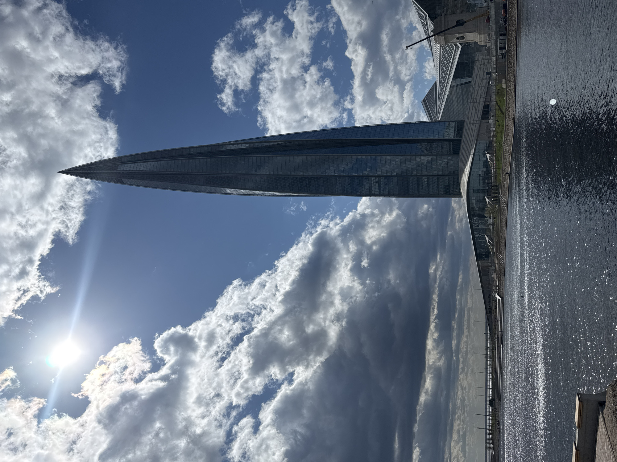 The image depicts the Lakhta Center skyscraper in Saint Petersburg, Russia. The building's pointed, needle-like silhouette rises against a blue sky filled with white and grey clouds. A body of water, likely the Gulf of Finland, is visible at the bottom of the frame. The sun is prominent in the sky, creating a lens flare effect. Construction activities are evident near the base of the Lakhta Center, involving cranes and other building structures. A pier or promenade runs alongside the water, with evenly spaced light posts lining it. The water surface reflects the sunlight and the buildings.