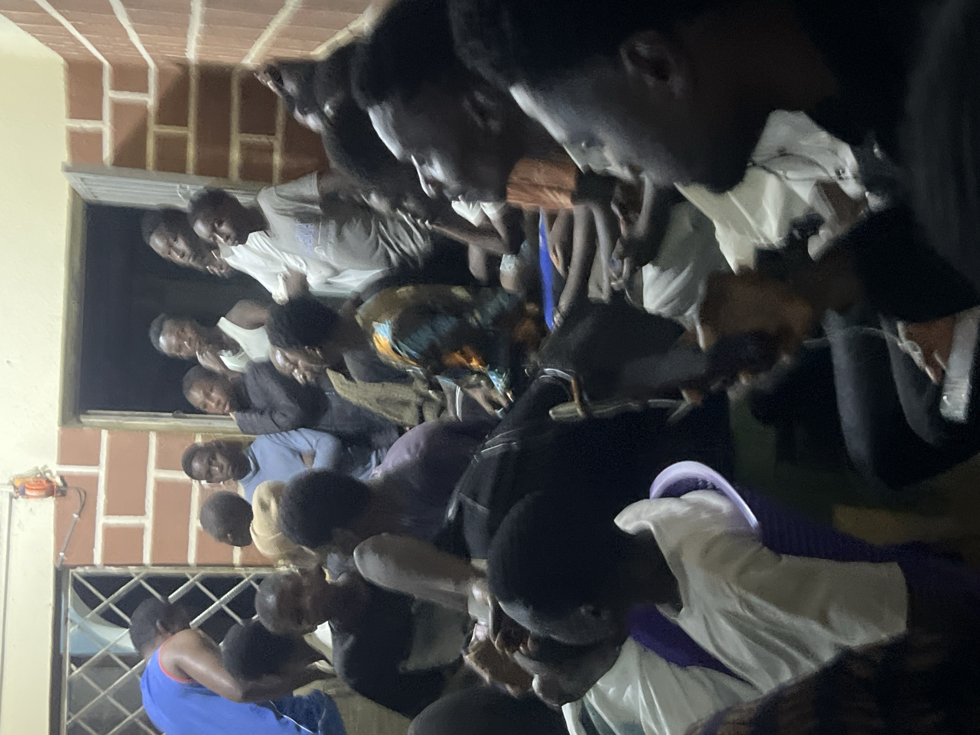This image depicts a gathering of people, predominantly young men and boys, in what appears to be an indoor setting with brick walls and a doorway. The individuals are standing and seated, with many looking towards a focal point outside the frame. The lighting suggests it is either evening or a dimly lit indoor space. Some individuals are holding mobile phones, and one person is holding what appears to be a Bible. The clothing styles suggest a casual and perhaps modest attire. The environment suggests a community or religious gathering. The image is likely taken in Jos, Nigeria, a city known for its diverse population and vibrant community life.