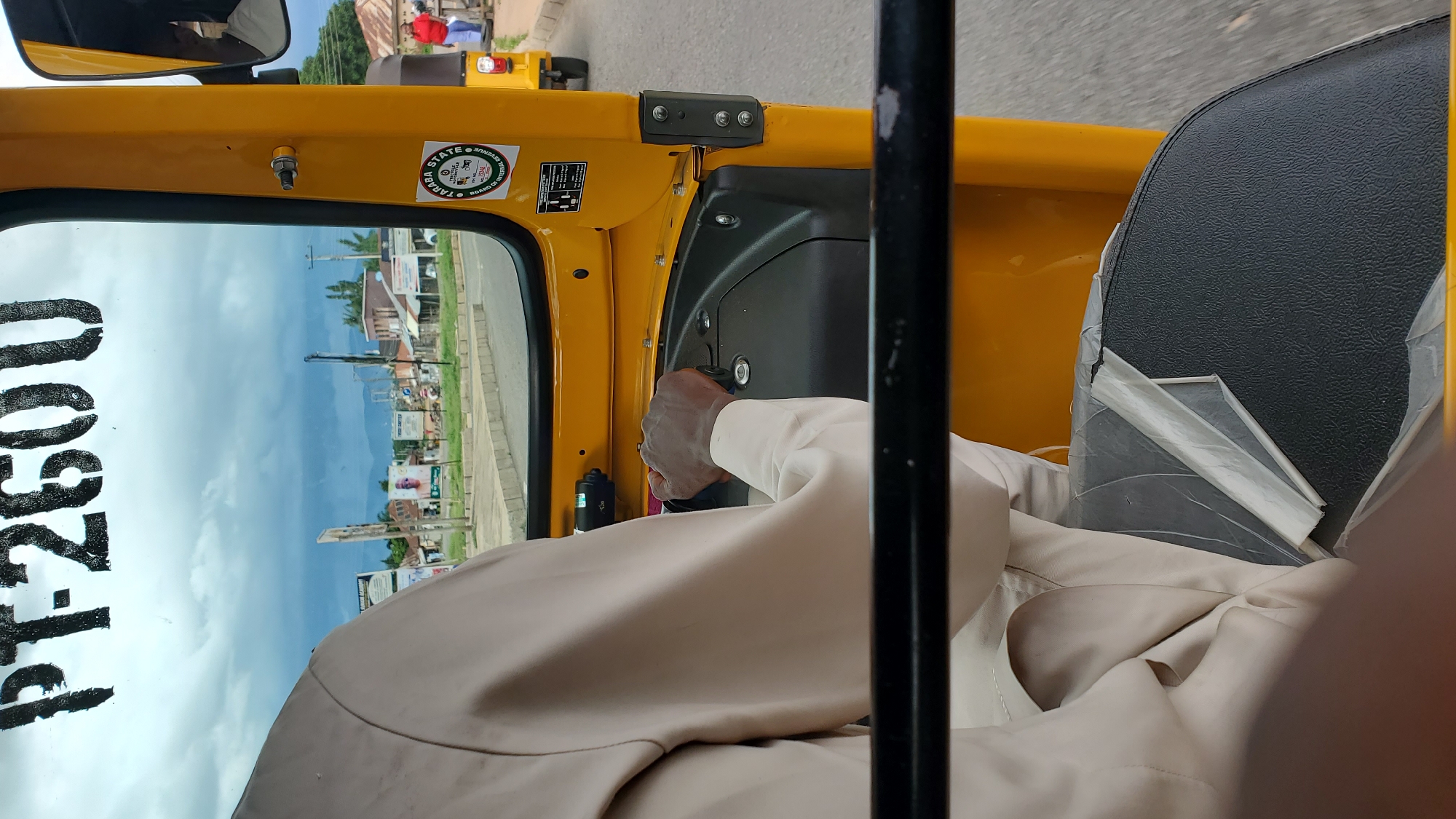 The image is a close-up view from inside a yellow auto-rickshaw, looking forward. The driver's hands, wearing a light-colored sleeve, are visible gripping the steering mechanism. The vehicle appears to be in motion on a paved road. Through the windshield, a street scene of Jalingo, Nigeria, is visible. Buildings, signs, and scattered vegetation line the road. The sky is overcast with dark clouds. A sticker labeled "TARABA STATE" is affixed to the yellow interior of the auto-rickshaw. Another auto-rickshaw is visible in the distance, as is a person wearing a red top.