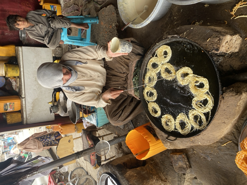 An image captured in Kandahar, Afghanistan, depicts a woman preparing a fried sweet snack, likely Jalebi, within a rustic storefront or stall.
The primary subject, a woman, is seated on the ground in the foreground, wearing a light-colored long-sleeved top, dark brown pants, and a light grey headscarf with a patterned band. She holds a metal tool in her right hand, manipulating multiple circular, golden-yellow items frying in a large, circular black pan filled with oil, which rests on a makeshift mud or brick stove structure. In her left hand, she holds a white cup. To her right, a white plastic tub contains a liquid batter. An orange plastic container sits to her left. Partially visible in the bottom left is a plate of finished, golden-orange sweets.
In the mid-background, a young boy in a light-colored traditional outfit sits barefoot on a blue plastic stool, observing the scene. Behind him, shelves are stocked with various items, including multiple yellow plastic jerrycans, other colored containers, and a white refrigerator/freezer unit. A gas cylinder and metal cooking vessels are also visible.
The scene opens to a street in the background, where a woman in a light brown burqa over a patterned dress walks away from the camera. Other individuals and motorcycles are visible further down the bustling street, indicating an urban market environment. The floor of the stall is packed dirt.