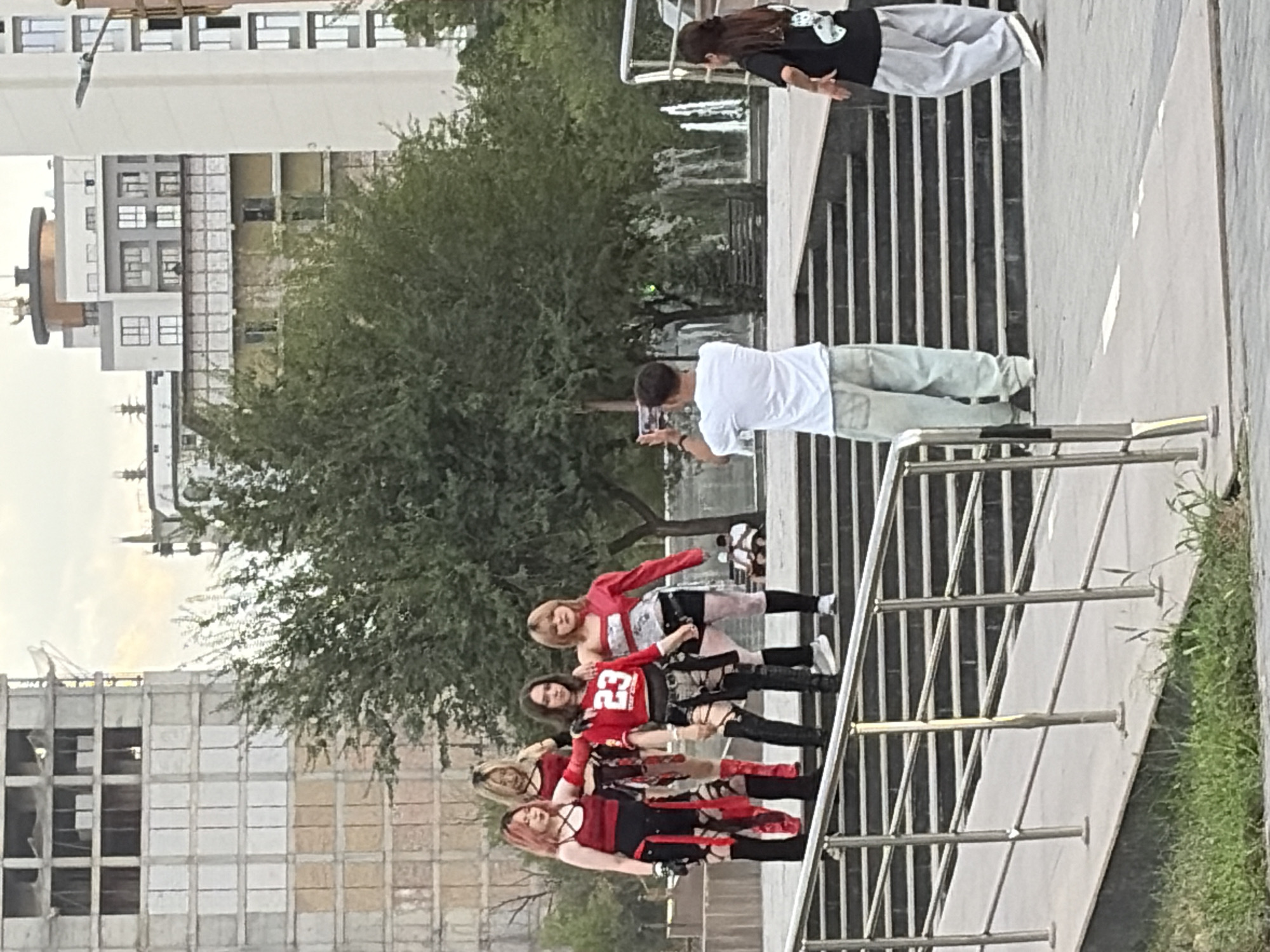 In an outdoor urban environment, a group of four young women dressed in matching red and black outfits, possibly a dance group or performers, pose for a photograph on a set of outdoor stairs. One woman in the center wears a red top with the number "23" prominently displayed. To their right, a young man in a white t-shirt and baggy pants holds up a smartphone, appearing to take a picture of the women. Further to the right and slightly behind him, a young woman in a black t-shirt and baggy pants stands facing away from the camera. In the background, several modern buildings are visible, including one under construction. Trees and a fountain are also present in the scene. The overall impression is a candid moment of urban life, with individuals engaged in recreational or artistic activities.